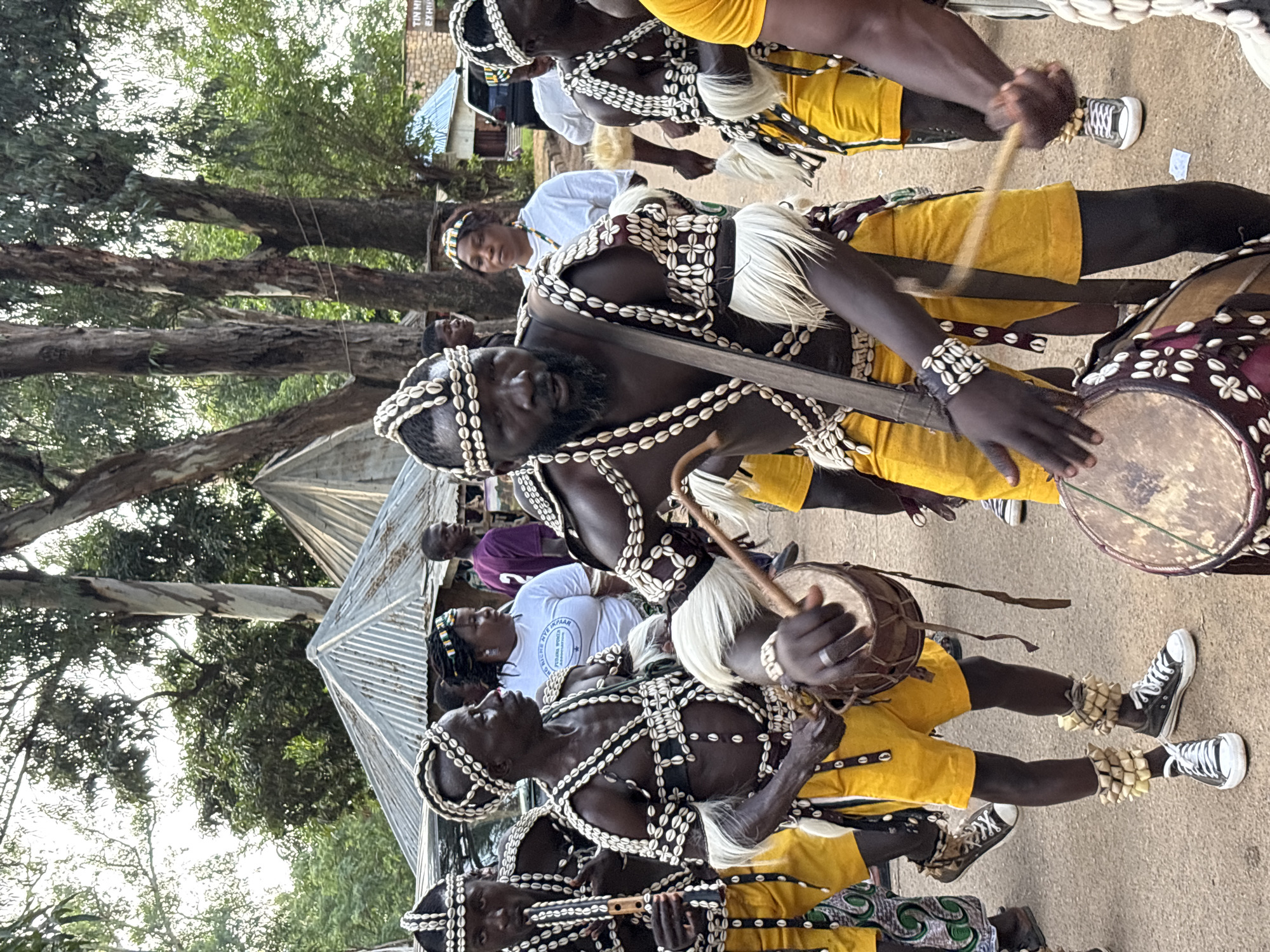 In an outdoor scene in Jos, Nigeria, a group of people, primarily men in traditional attire, are gathered. The men are adorned with headdresses and chest coverings made of white shells and beads, paired with yellow shorts. Several individuals are actively playing drums and one is playing a flute, suggesting a musical performance or cultural event. In the background, other individuals are visible, some in casual clothing and others in similar traditional wear. Tents and buildings are present, indicating a village or community setting. The overall atmosphere appears to be celebratory or ceremonial.