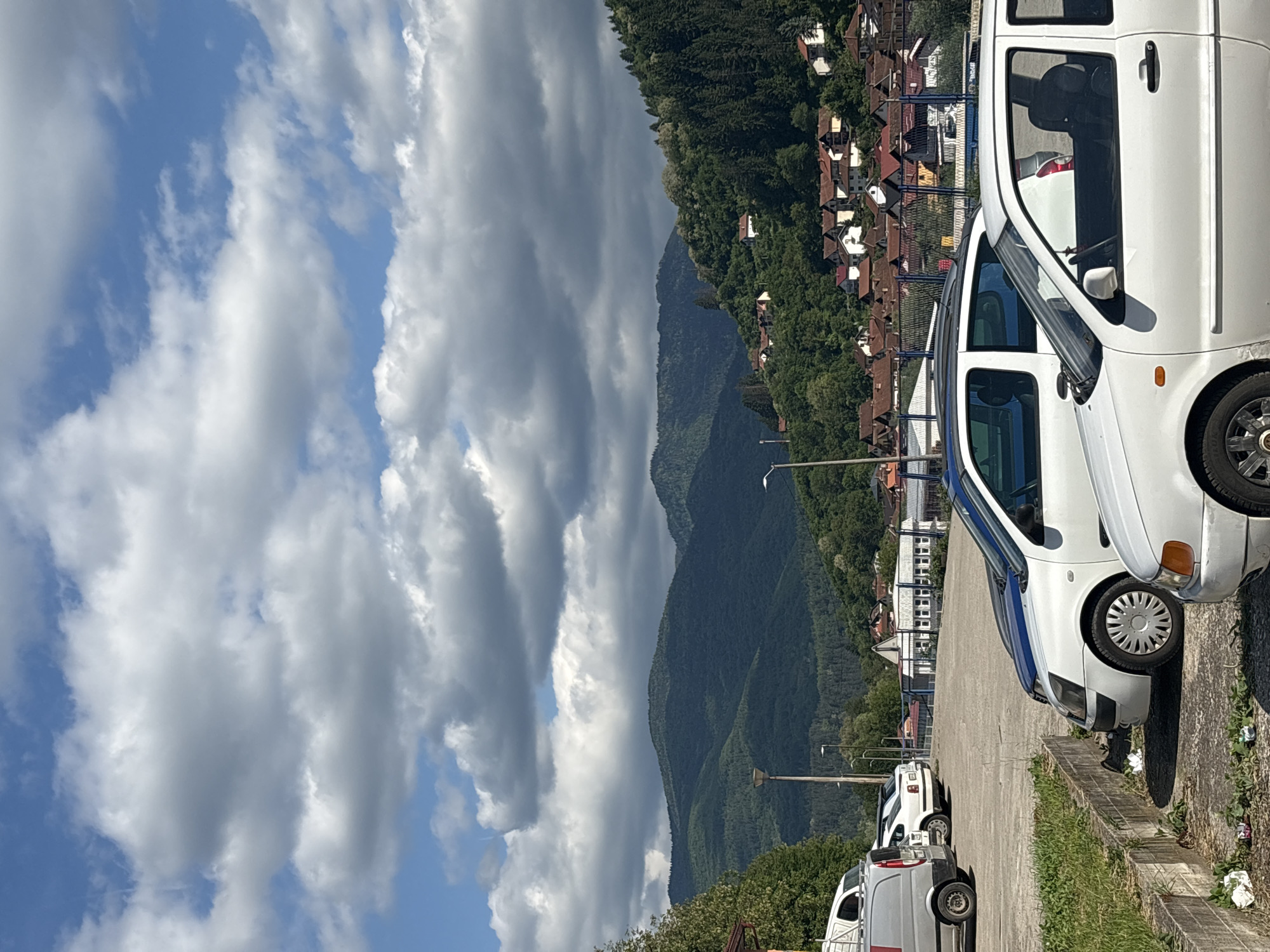 The image shows a view in Foča, Bosnia and Herzegovina. A paved parking lot is in the foreground, with several white vans parked along its edge. A low concrete barrier separates the parking lot from a grassy area. Beyond the parking lot, a mountain range covered in dense green forest rises in the background. Houses with red roofs are visible nestled among the trees. The sky is filled with partly cloudy blue sky. The lighting is bright, suggesting a sunny day.