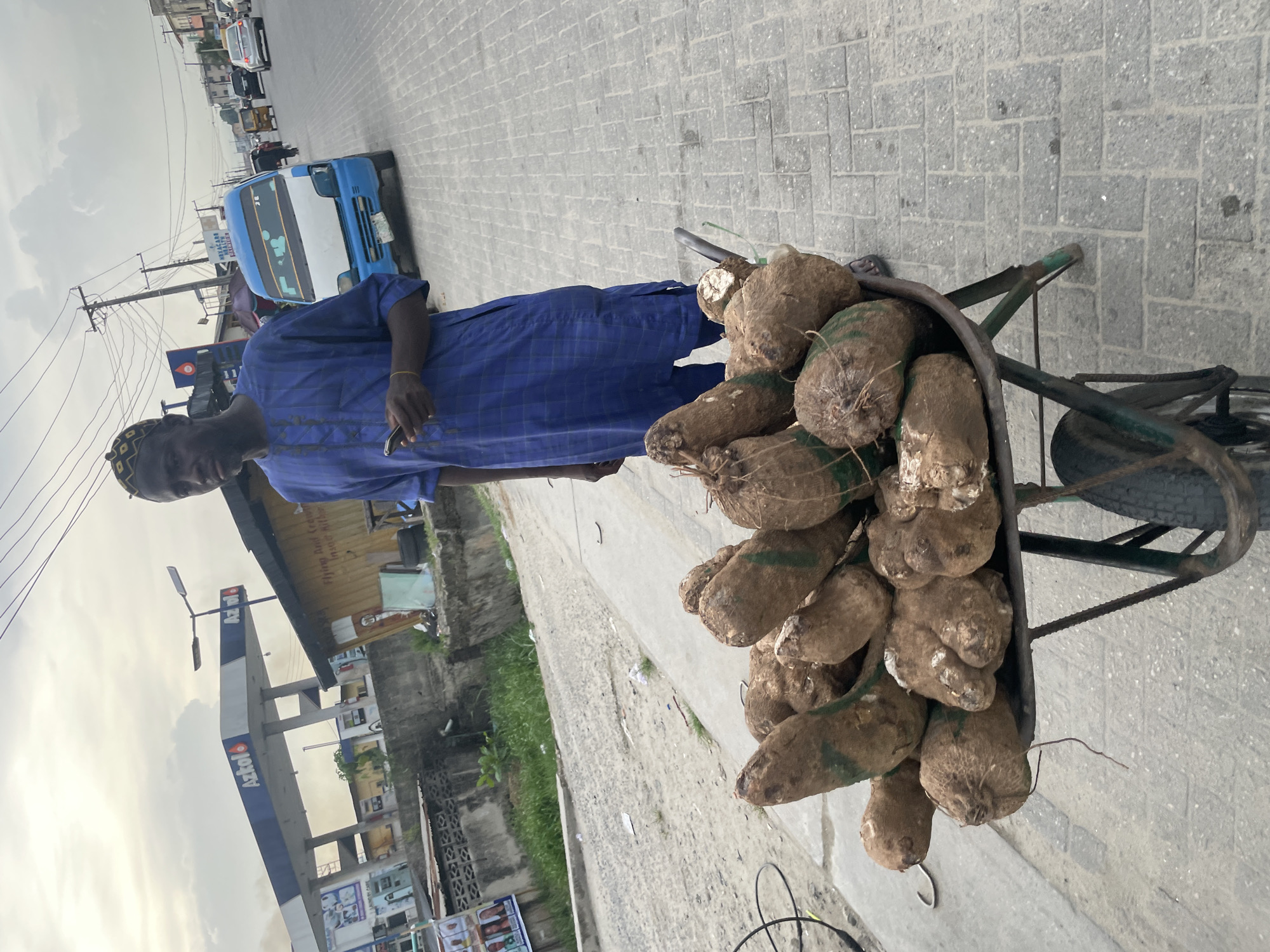 This media file captures an outdoor street scene in Ado, Nigeria, featuring a yam vendor and his handcart. The image is rotated 90 degrees clockwise.
In the foreground, a man of African descent stands beside a metal handcart overflowing with large, brown yams. Some of the yams are marked with green bands or ties. The man is dressed in a blue, patterned long-sleeved tunic and a matching cap. He holds a small, metallic object in his right hand and wears a light-colored bracelet on his left wrist. His expression is neutral.
The setting is a paved street and sidewalk made of grey interlocking blocks. Behind the man, a busy road is visible with several vehicles. A prominent blue and white minibus, characteristic of Nigerian public transport, is clearly seen. Text partially visible on the bus includes "WELCOM" and "COLLEGE." Further down the road, other cars and smaller vehicles are present.
To the man's left, a fuel station with a tall blue and white sign displays the brand "Aztoll" (or "Azrol") and "MOTORS." Below it, a smaller white sign indicates "Filling And Cream" and "laundry." Utility poles with numerous overhead wires are scattered throughout the scene. Buildings, including residential structures and commercial establishments, line the street.
The sky is overcast, indicating a daytime setting, likely late morning or afternoon, with diffused lighting. The image provides a candid glimpse into a typical street commerce activity in an urban Nigerian environment.