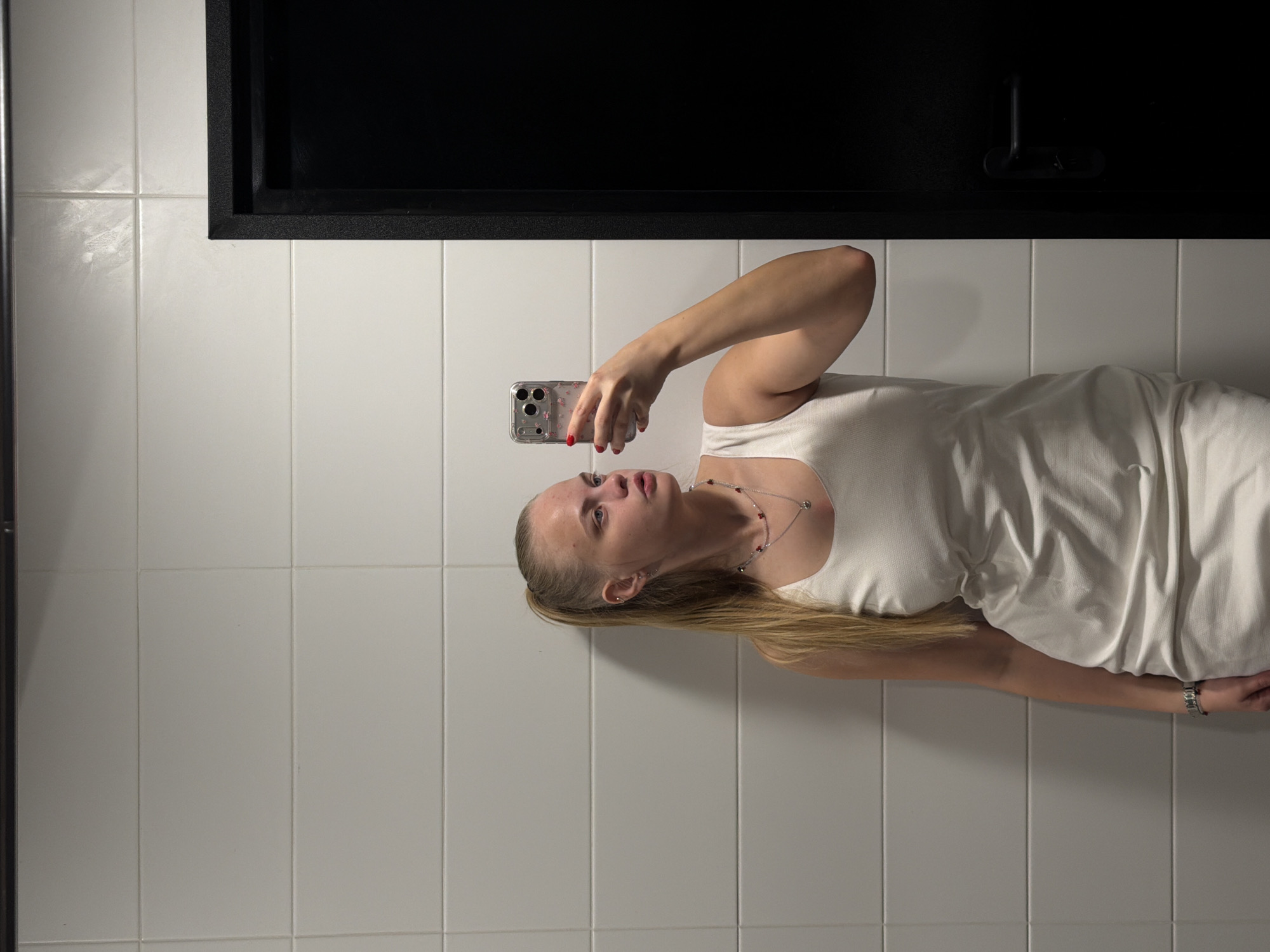 A young woman is holding a smartphone and posing for a photograph in a bathroom. The background consists of white square tiles. Above her is a rectangular dark-colored recess or storage compartment in the tiled wall. The woman has blonde hair, is wearing a white top and a necklace, and has red nail polish. No specific information suggests the location is Izhevsk, Russia.