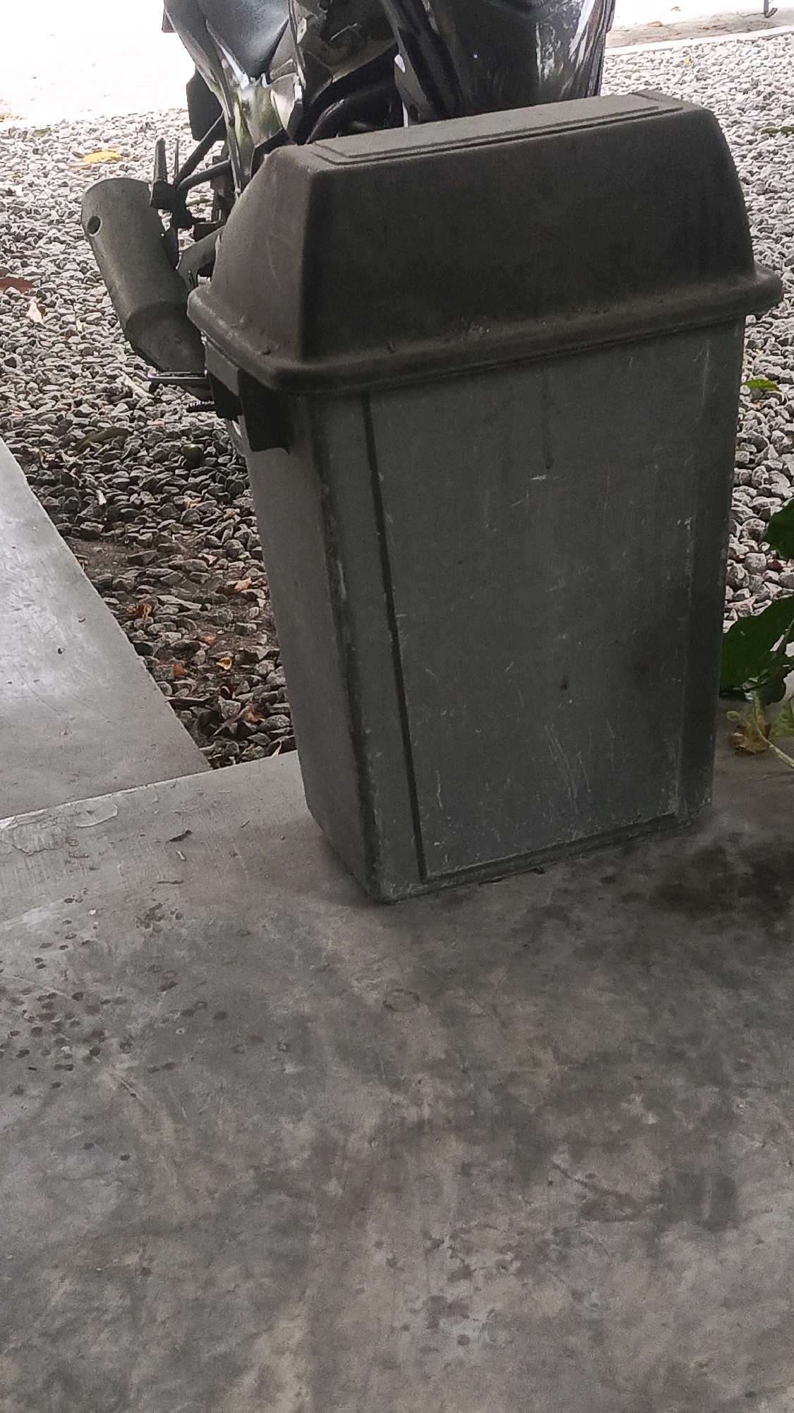 A dark gray trash bin with a black lid sits on a concrete surface. Behind the trash bin, a motorcycle is partially visible, with its exhaust pipe extending towards the left. The ground surrounding the trash bin and motorcycle is covered in gravel and scattered leaves. A concrete ledge runs along the left side of the image, leading away from the viewer. The overall scene appears to be outdoors.