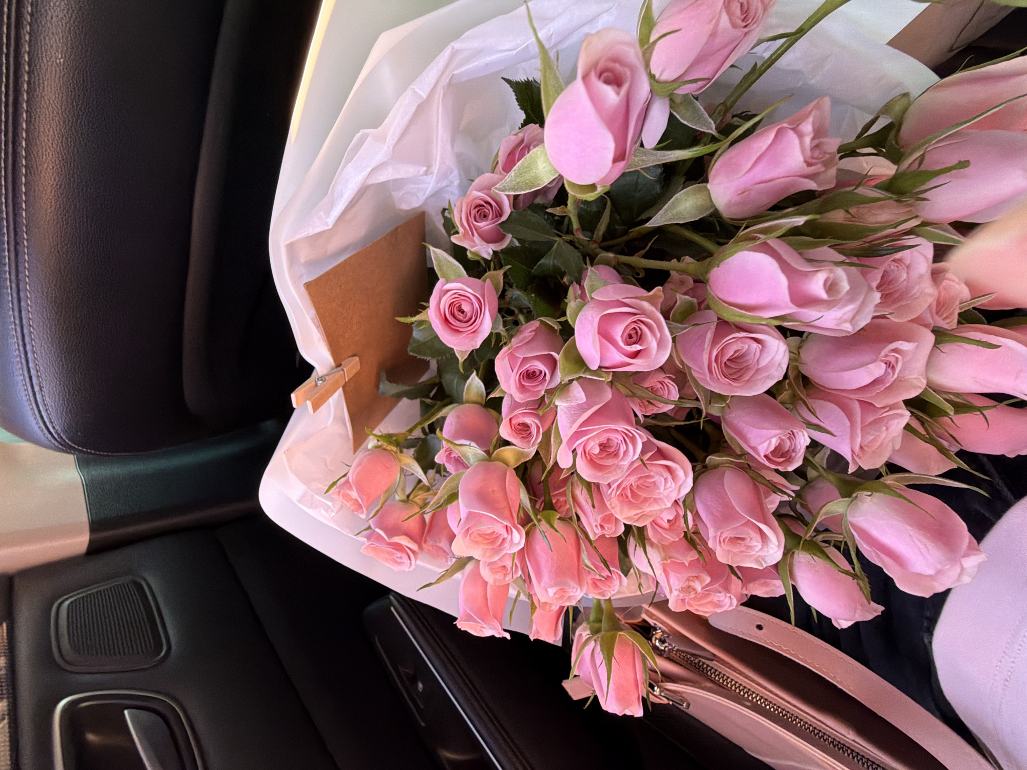 A bouquet of pink roses is placed on the passenger seat of a car, partially wrapped in white paper. A small brown card is attached to the wrapping with a wooden clothespin. To the left of the roses is a pale pink purse with a zippered compartment. The interior of the car is visible, featuring black leather upholstery on the door and seat. The scene is static, and no individuals are present.