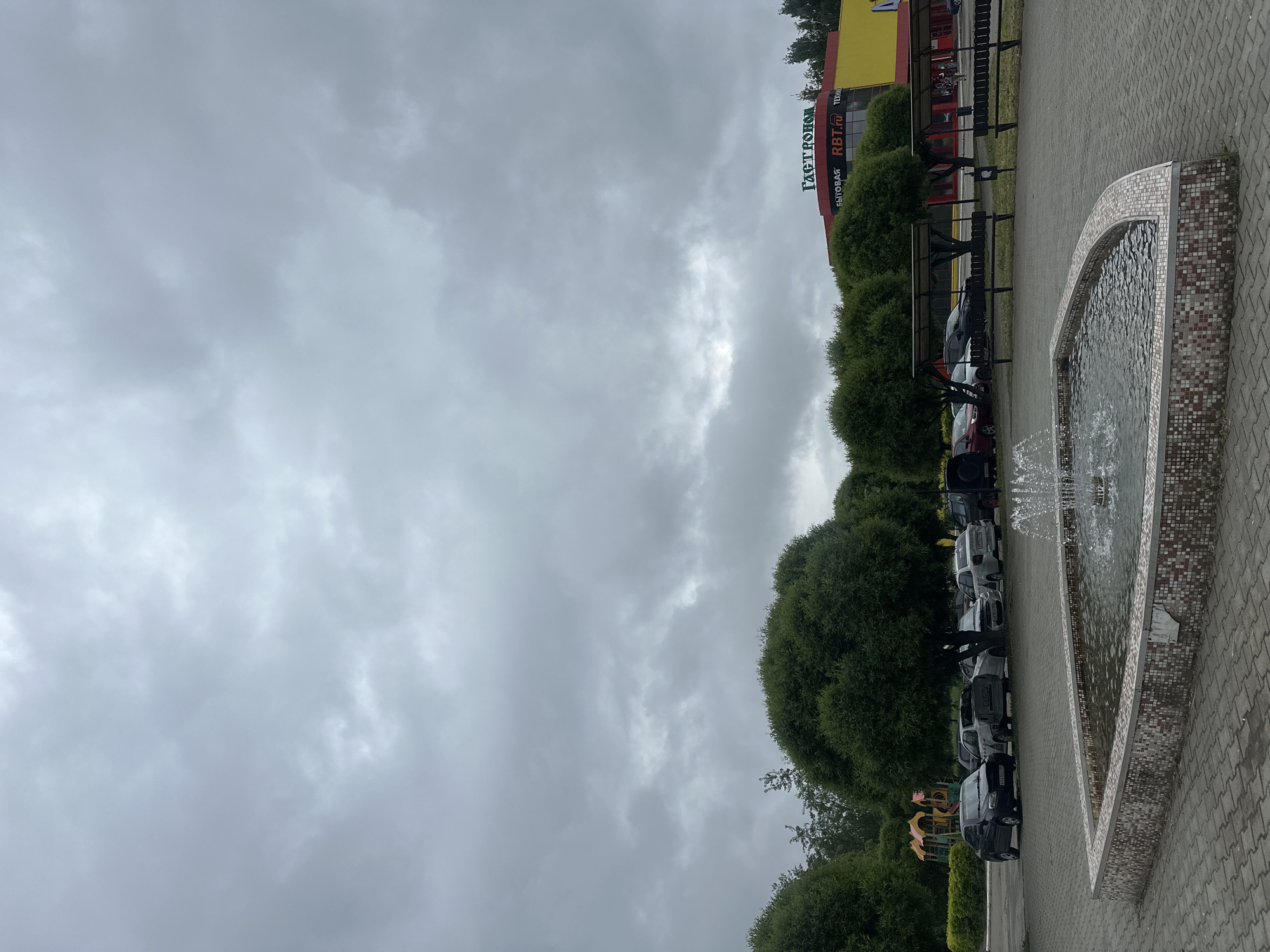 The image shows a cloudy outdoor scene in what appears to be the city of Chaikovsky, Russia. A fountain is in the foreground, with water spraying upwards. Behind the fountain is a row of parked cars, separated from the building by a line of trimmed bushes. Further back is a yellow and red building with lettering in Russian, reading "ГАСТРОНОМ". The weather appears overcast, with a gray sky.