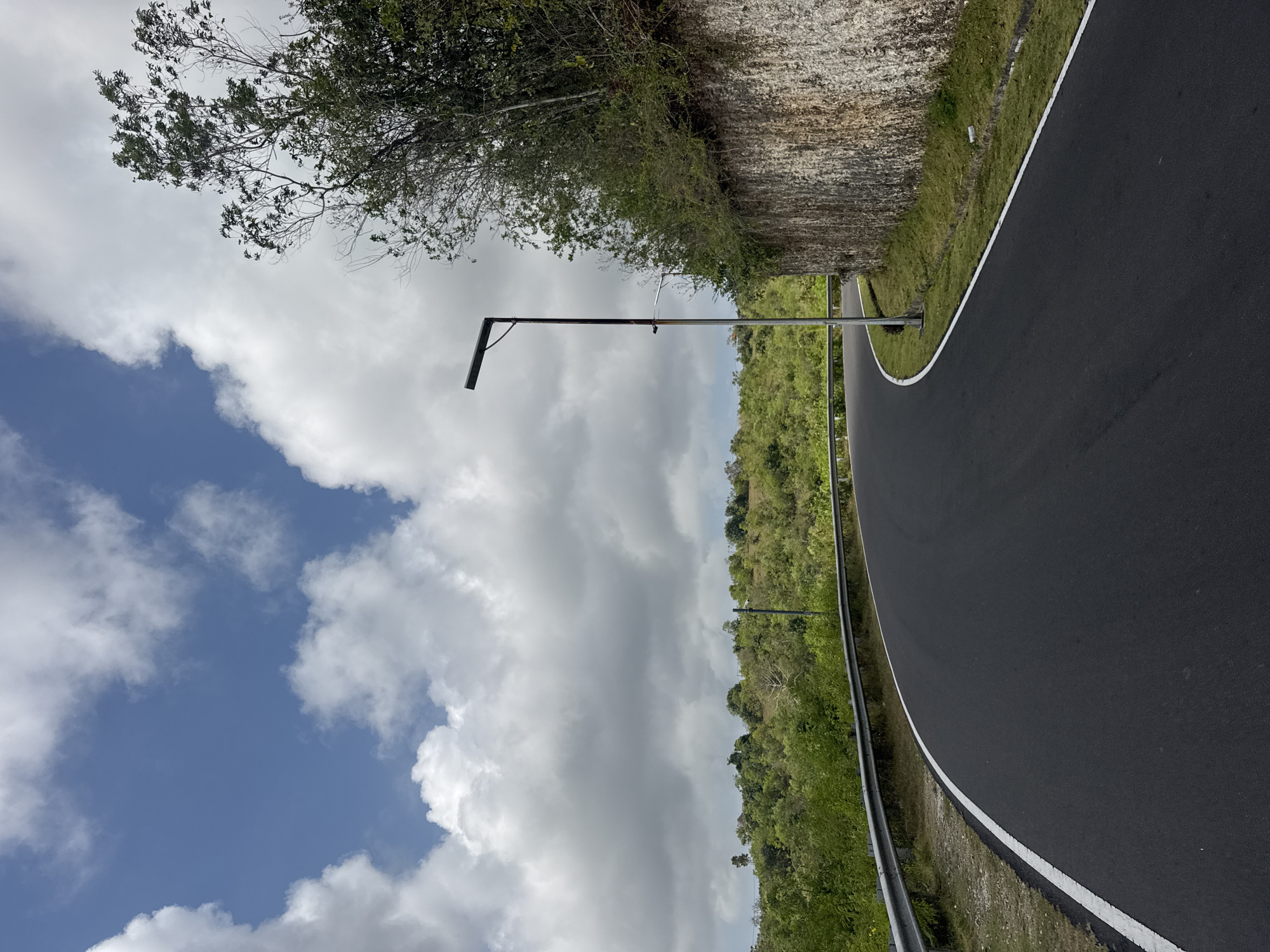 A paved road curves through a lush, green landscape under a partly cloudy sky. The road surface is dark asphalt with a white line marking its edge. On the left, a metal guardrail separates the road from a vegetated slope. On the right, a rough, light-colored stone wall rises, topped with dense green foliage and trees. A tall, slender lamppost with a dark fixture stands near the road, its base close to the stone wall. In the distance, rolling hills covered in trees and vegetation stretch towards the horizon. The sky is a vibrant blue, punctuated by fluffy white clouds. This scene appears to be on a well-maintained road in a hilly, tropical environment, potentially within the city of Kuta Selatan, Indonesia, given the vegetation and landscape. There are no people or animals visible in the image, and no discernible actions or interactions taking place.