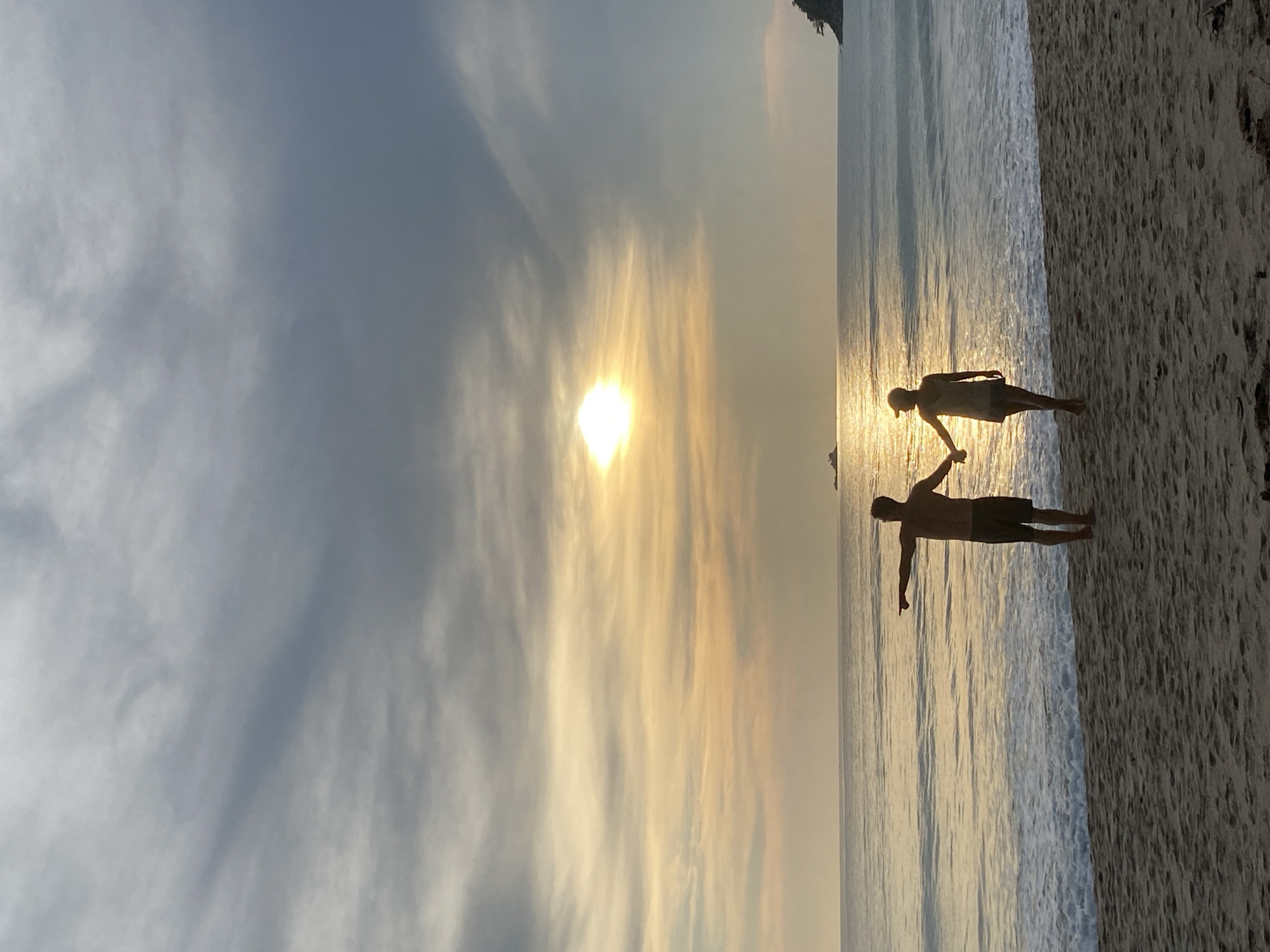 A silhouette of two people is visible on the beach in Choeng Thale, Thailand, at sunset. The sky is filled with clouds, with the sun shining through a break in the clouds. The ocean is calm, with small waves lapping at the shore. The sand is light in color. One person is standing with arms outstretched, while the other is holding hands with the first person and holding a neutral arm position.
