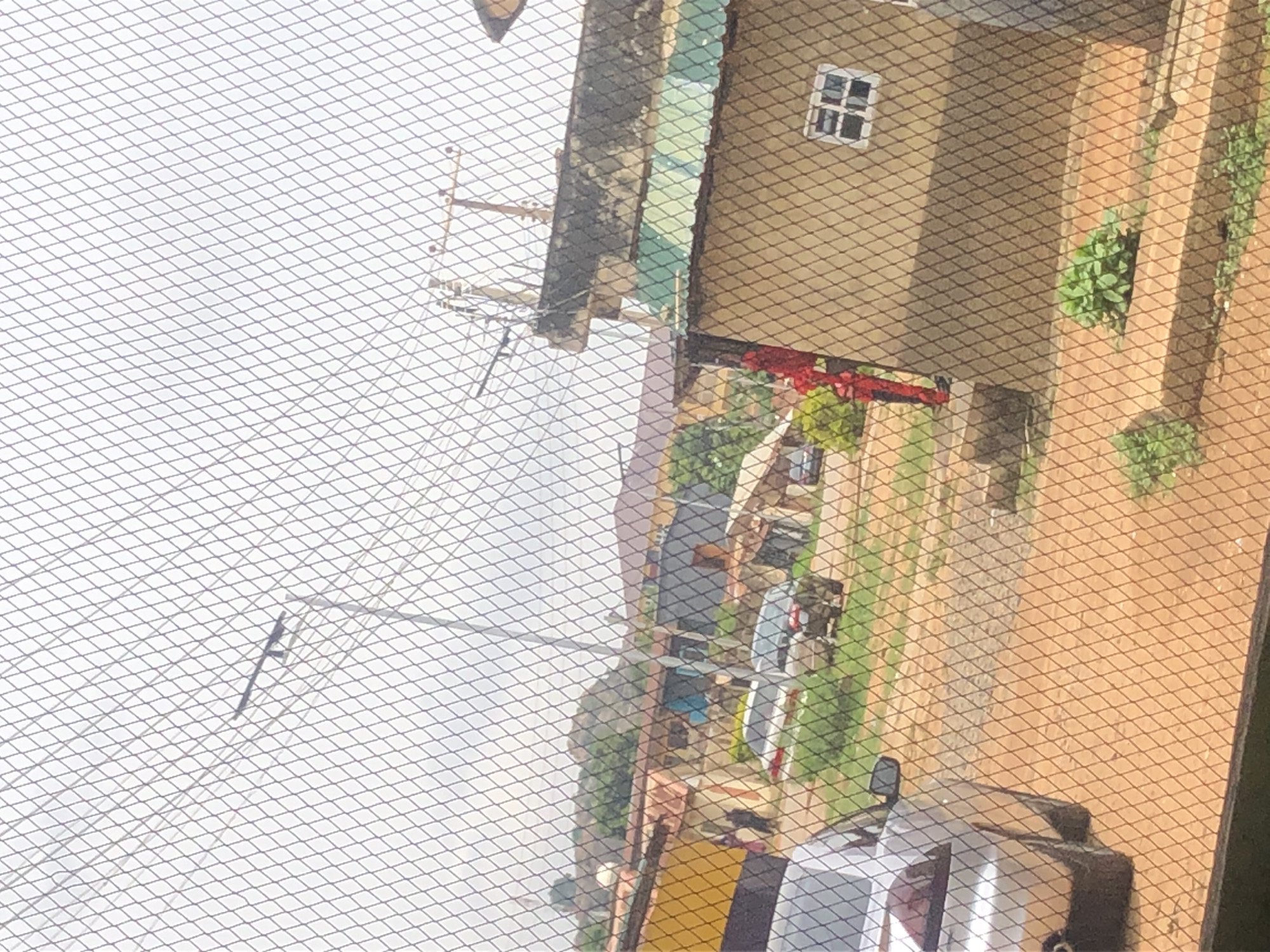 A security fence with a diamond mesh pattern obstructs the view of a street scene in a developing urban environment. In the foreground, a silver car is partially visible on the left side, with its rear end facing the viewer. The car is parked on a dirt or gravel surface. Behind the fence, a road or pathway with a reddish-brown, dusty surface stretches into the background.
On the right side of the image, a tan-colored building with a dark, weathered roof is prominent. The building has a small, square window with multiple panes and a white frame. A person dressed in a red uniform is standing near the entrance of this building. Further back, several other buildings with various roofing materials, including corrugated metal and darker shingles, are visible. Greenery, including bushes and trees, is scattered throughout the scene, suggesting a somewhat unkempt or natural landscape encroaching on the developed area. The sky is overcast with a light gray hue. Power lines and poles are visible in the background, indicating the presence of infrastructure. The overall impression is of a transitional or semi-developed area.