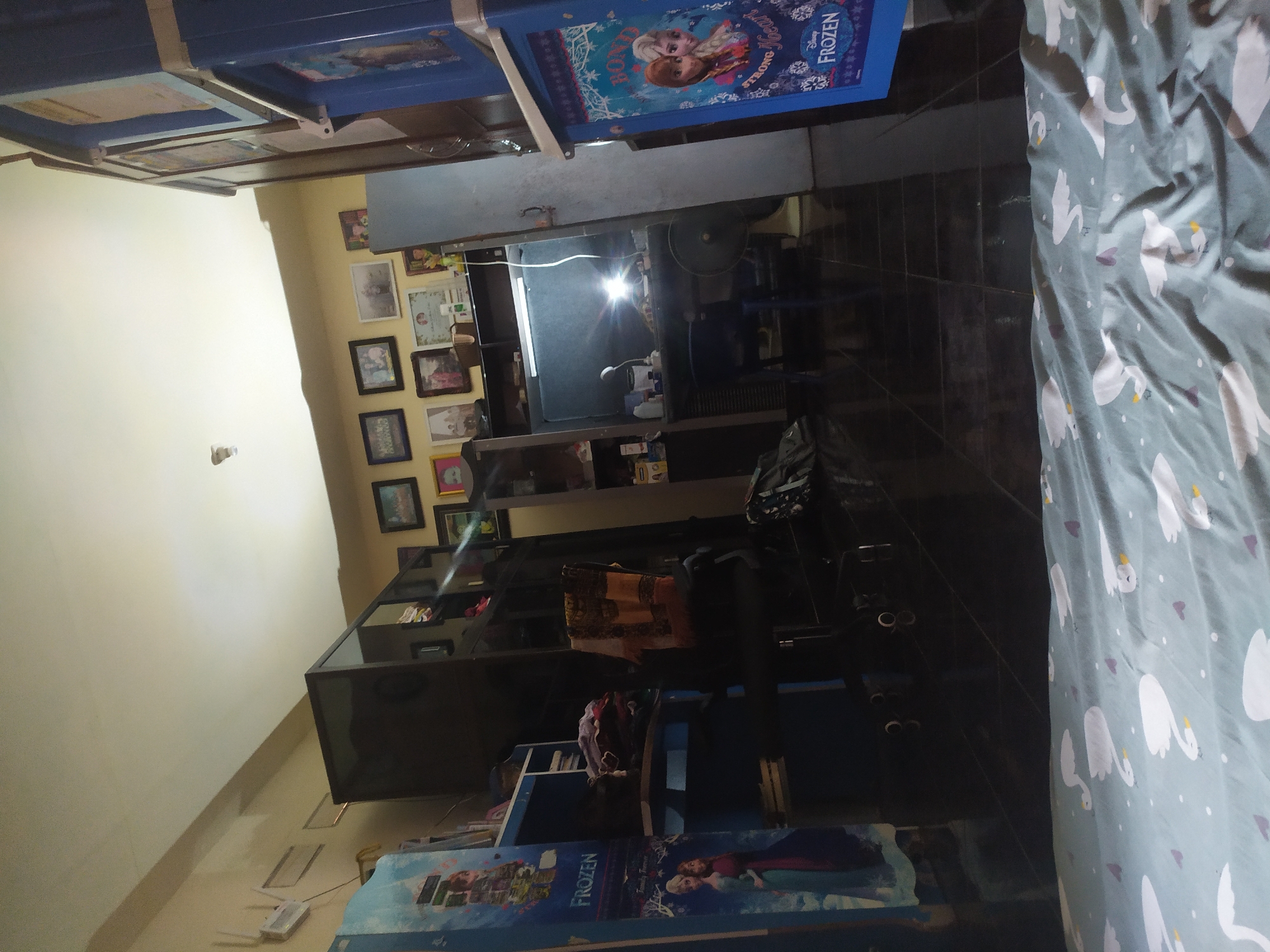 The image depicts a dimly lit room with tiled flooring. On the left, a blue cabinet with "FROZEN" branding is visible, featuring characters from the movie. Next to it, a dark cabinet unit is present. In the center, a desk with a lamp and a fan is set up. Behind the desk, shelves are lined with framed pictures and various items. To the right, another blue cabinet unit, also displaying "FROZEN" imagery, is situated. The walls are painted a light yellow. The ceiling has visible light fixtures. The overall impression is of a bedroom or living space, potentially in a residence in Kragilan, Indonesia, given the context.