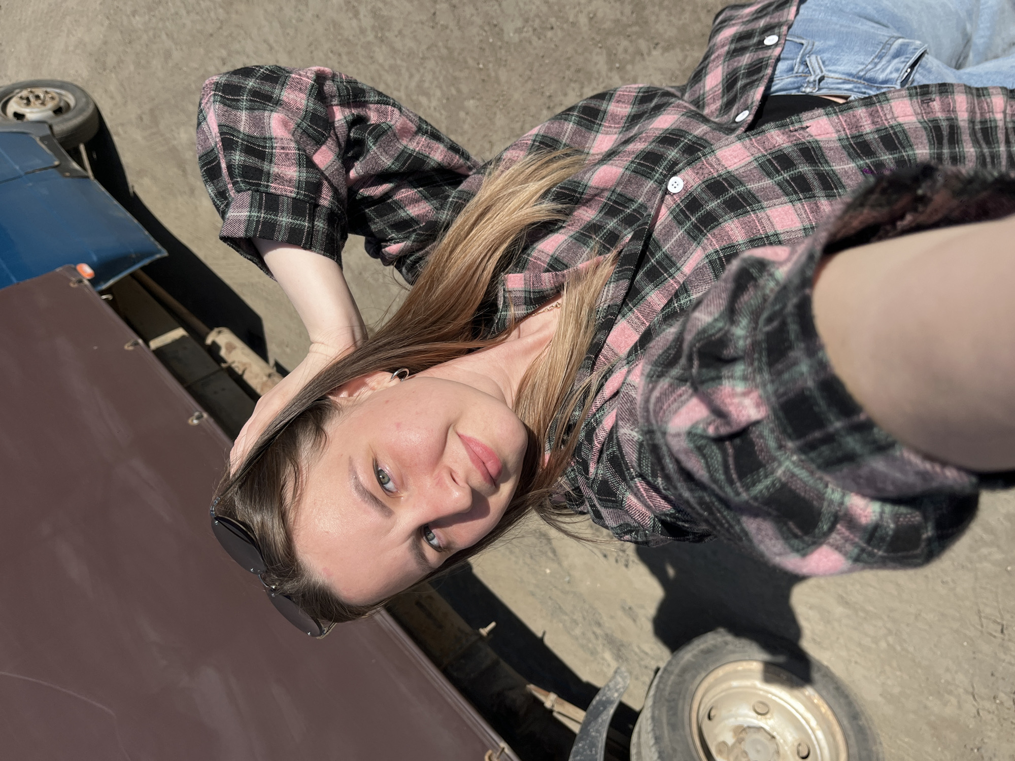 A light-skinned female lies on her back on a concrete surface outdoors. She has shoulder-length, blonde-brown hair and wears a pink and black plaid button-down shirt over blue jeans. Sunglasses rest on her head. A brown, possibly metal, object is to her left. A blue and silver vehicle, possibly a trailer, is on the upper left. The ground is dry and light-colored. The location is undetermined and cannot be identified as the city of Izhevsk, Russia, from the visual information provided.