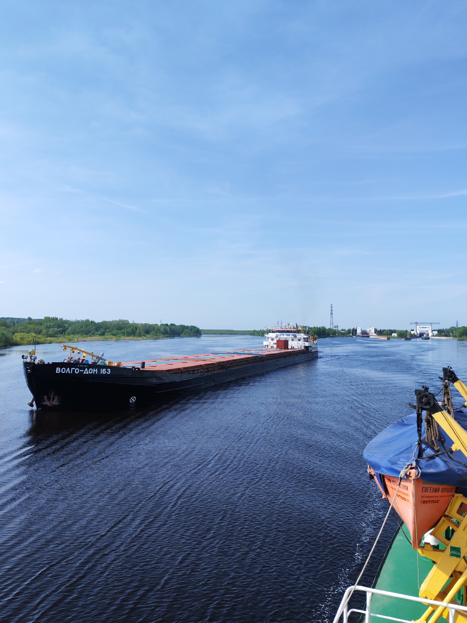 The scene depicts a wide river view with a barge named "BOJTO-DOH 163" prominently featured in the left half of the frame. The barge is black with white Cyrillic lettering. Its deck is visible, appearing dark red or brown. The river's surface shows rippling water, moving towards the viewer.
In the background, the riverbanks are lined with green vegetation, and a distant structure, possibly industrial or related to the river infrastructure, can be seen. On the right side of the frame, a portion of another vessel is visible, with a bright orange lifeboat covered by a blue tarp being the most noticeable element. The sky is a pale blue with scattered thin clouds.
Based on the available information and the barge's name Волго-Дон, which refers to the Volga-Don Canal, it is reasonable to infer that the image was taken on a river connected to the Volga-Don waterway. While without further corroborating details it is not possible to definitively place the image in Нифантово, Russia, it is plausible given the geography and the nature of the river transport depicted. The scene is absent of human presence beyond the presumed crews of the vessels.