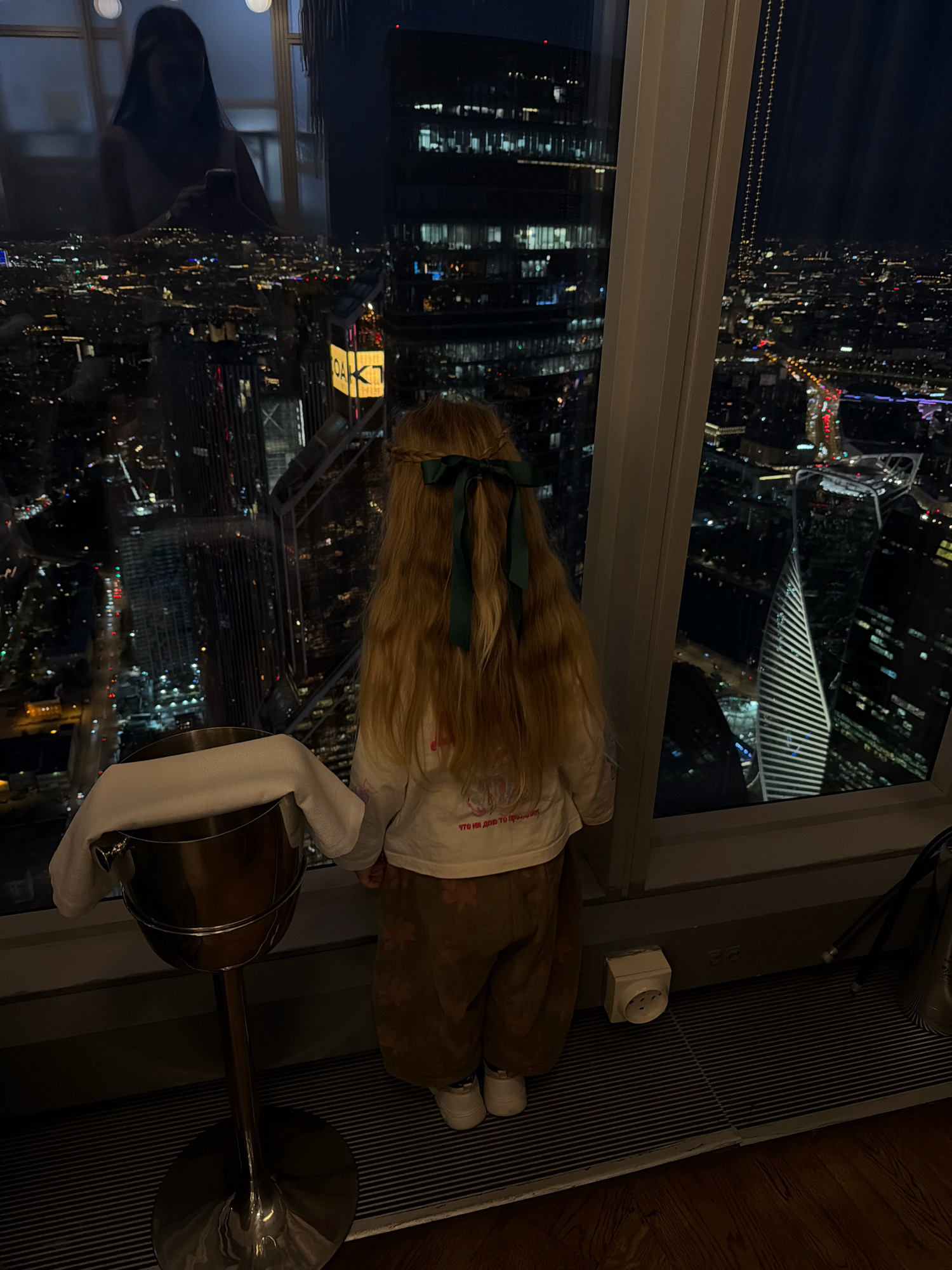 A child with long blonde hair tied with a dark green ribbon stands facing away from the camera, looking out a large window at a city skyline at night. The child is wearing a white long-sleeved shirt with graphics and tan pants with a floral pattern. To the left of the child is a silver champagne bucket with a white cloth draped over it. The window overlooks a sprawling cityscape with numerous illuminated buildings and lights. In the reflection of the window, a person holding a phone can be seen. The scene appears to be set in an indoor environment, possibly a restaurant or hotel, given the view and the presence of the champagne bucket. The floor beneath the child and bucket is a dark ribbed mat, with wooden flooring visible to the right. A white electrical outlet is mounted on the wall. The image is captured at an angle, with the city lights providing a dramatic backdrop to the foreground elements. The overall impression is one of quiet observation of the urban landscape from a high vantage point.