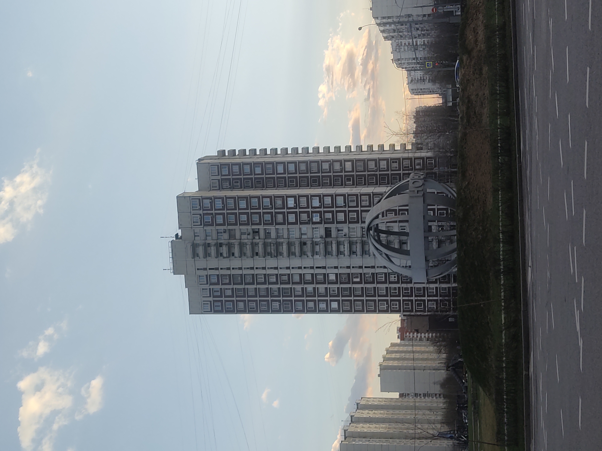The image shows a tall, square-windowed building adjacent to a multi-lane roadway in Moscow, Russia. The building's architecture appears to be Soviet-era. A large, spherical metal structure with inscriptions is positioned near the building. Another similar building is visible in the background. There is a median strip with vegetation between the roadway. The sky is clear with some clouds.
