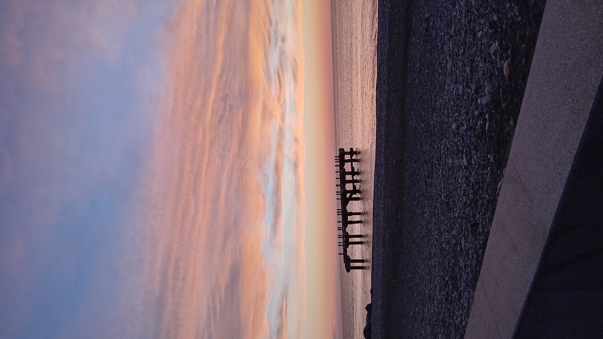 The image depicts a sunset at Batumi Beach in Batumi, Georgia. The sky displays hues of orange, pink, and blue. A calm sea reflects the colors of the sunset. A portion of a dark, rocky beach is visible on the right side of the image, next to a concrete structure. Old, wooden posts, remnants of a former pier, stand in the water.