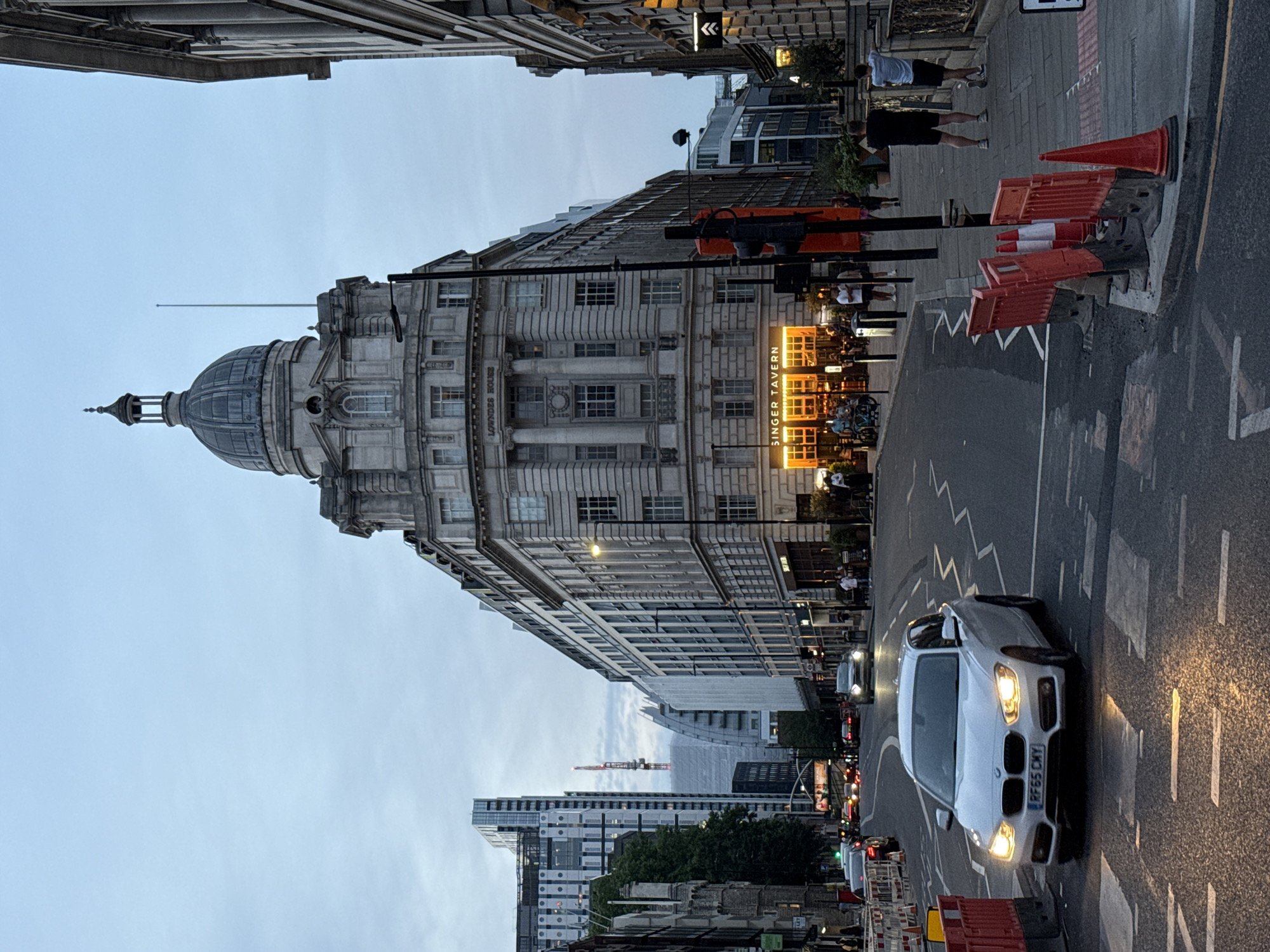 The image shows a street scene in London, United Kingdom, dominated by a large, ornate building with a domed roof, identifiable as Lowndes House. A white BMW with registration RF65 CXY drives on the right side of the road. The street includes orange and white traffic cones, pedestrians, a building called Singer Tavern, and a traffic light. In the distance are additional modern high-rise buildings and a construction crane. The sky is overcast.
