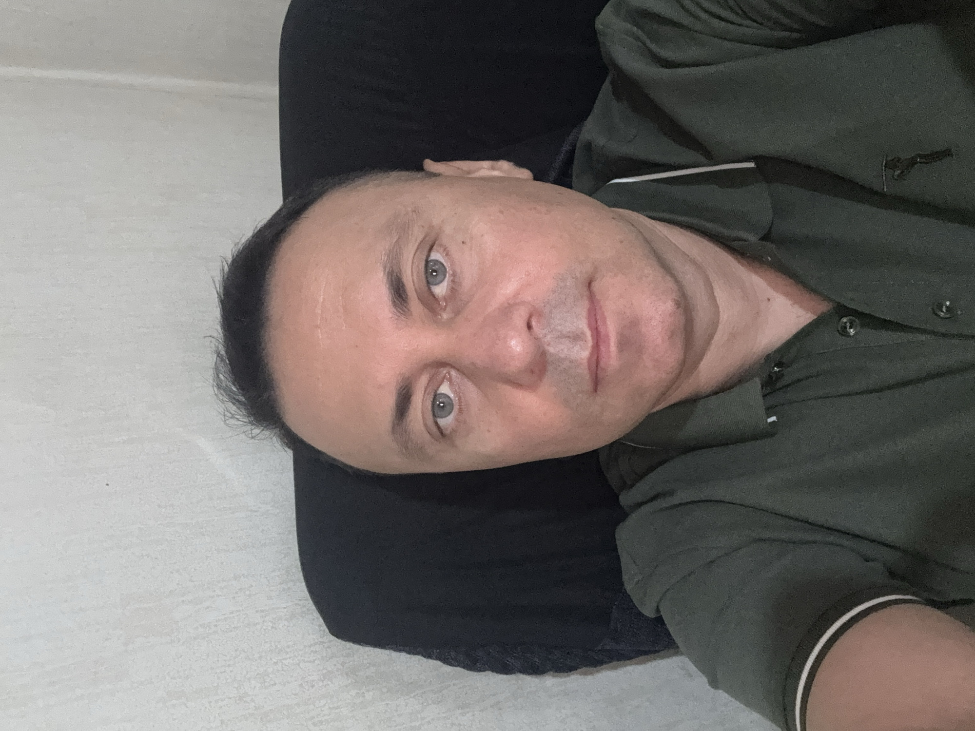 The image shows a man seated in a chair. He is looking directly at the camera and appears to be indoors. The background consists of a light-colored textured wall on the left and a dark object, possibly a chair or cushion, on the right. The man is wearing a dark green polo shirt with white trim on the collar and sleeves. There is a logo on the left side of his shirt. The overall lighting suggests an indoor setting, possibly a home or office. There is no information in the image to indicate the specific location in Tbilisi, Georgia.