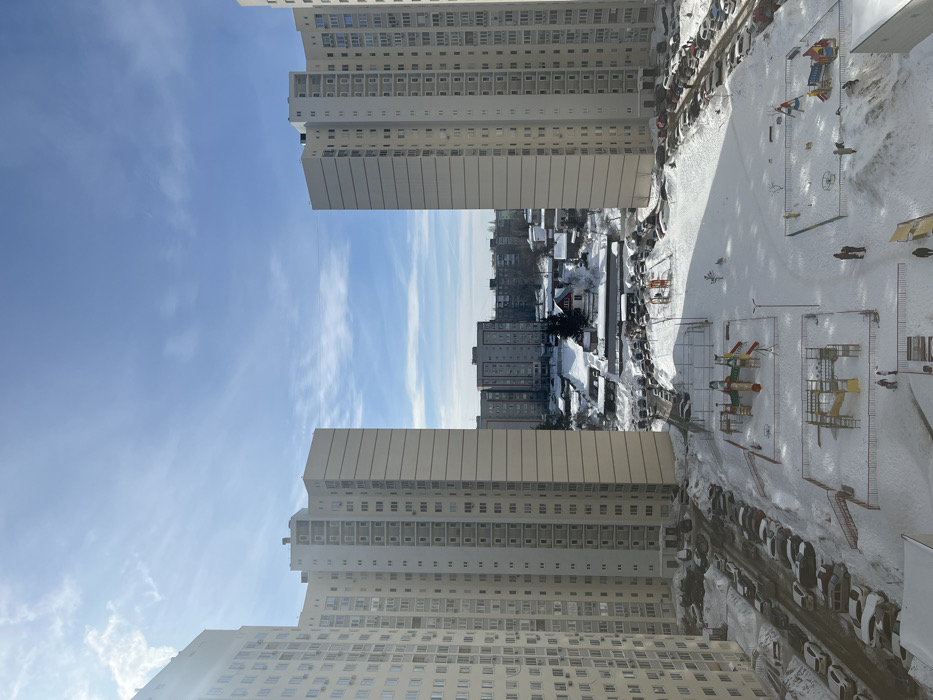 The image depicts a snow-covered residential urban landscape in Zvezda, Russia, observed from an elevated perspective during daytime under a bright blue sky with sparse clouds and a visible sun flare in the upper left. Two prominent, multi-story, light-colored (beige/off-white) residential buildings frame the scene, with the left building partially cropped. Both buildings feature numerous windows and integrated balconies.
The ground area between these structures is entirely covered in undisturbed white snow, revealing a large courtyard. Two distinct children's playgrounds, each enclosed by low fencing, are present in the snow. The left playground features a multi-colored (red, yellow, blue, green) climbing structure with slides. The right playground includes a smaller yellow-and-blue climbing structure, a circular merry-go-round, and another play element.
Multiple asphalt parking lots flank the central courtyard, filled with numerous parked vehicles partially covered in snow. Cleared vehicle tracks and pedestrian paths are visible, exposing dark pavement. Several individuals are present on the snow-covered ground: at least two adults and one child are near the right playground, and two individuals (one adult, one child) are walking on a cleared path in the lower-left portion of the image.
In the mid-background, a dense cityscape of additional multi-story apartment buildings is visible, interspersed with smaller structures. A distinctive small building with a red pitched roof and white walls stands centrally in the mid-background. A faint utility line stretches horizontally across the sky between the two dominant foreground buildings. Streetlights are present within the courtyard.