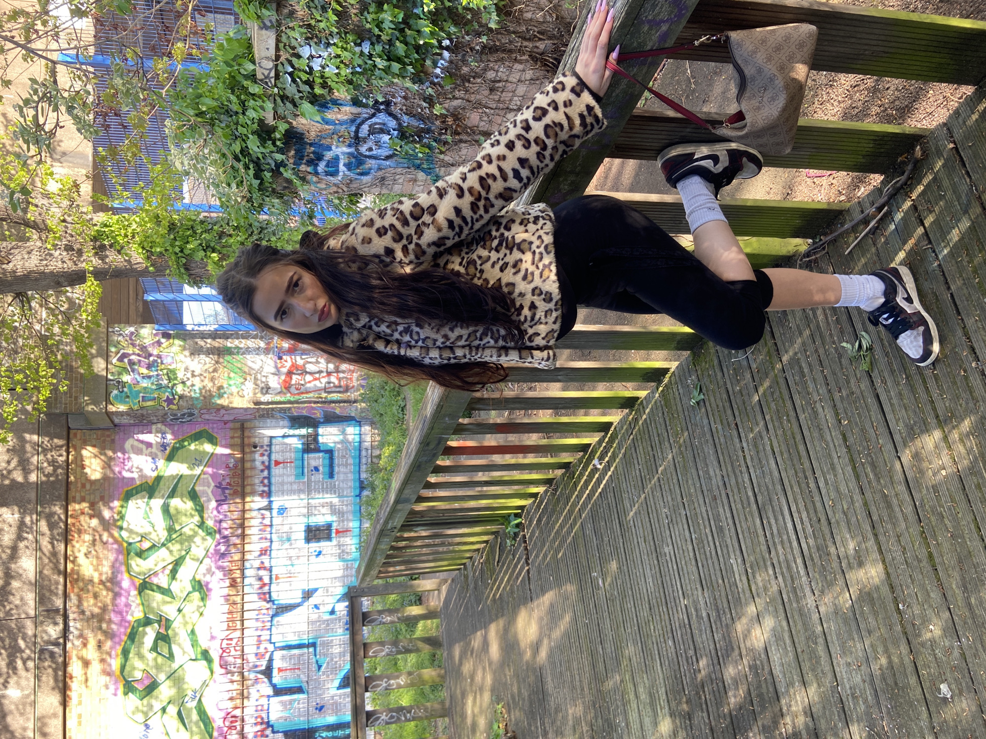 A woman is posing on a wooden walkway next to a brick wall covered in graffiti. The woman has long brown hair and is wearing a leopard-print jacket, black leggings, white socks, and black and white sneakers. She is holding onto a wooden railing with her left hand and is looking at the camera. Her right hand is near a purse hanging on the railing.
The graffiti on the brick wall includes various colors and designs. A tree and other foliage are present on the other side of the railing. The walkway is made of wooden planks and has a slightly worn appearance. The location is likely a public walkway in an urban environment. The location is unknown, and can not be identified from the image.