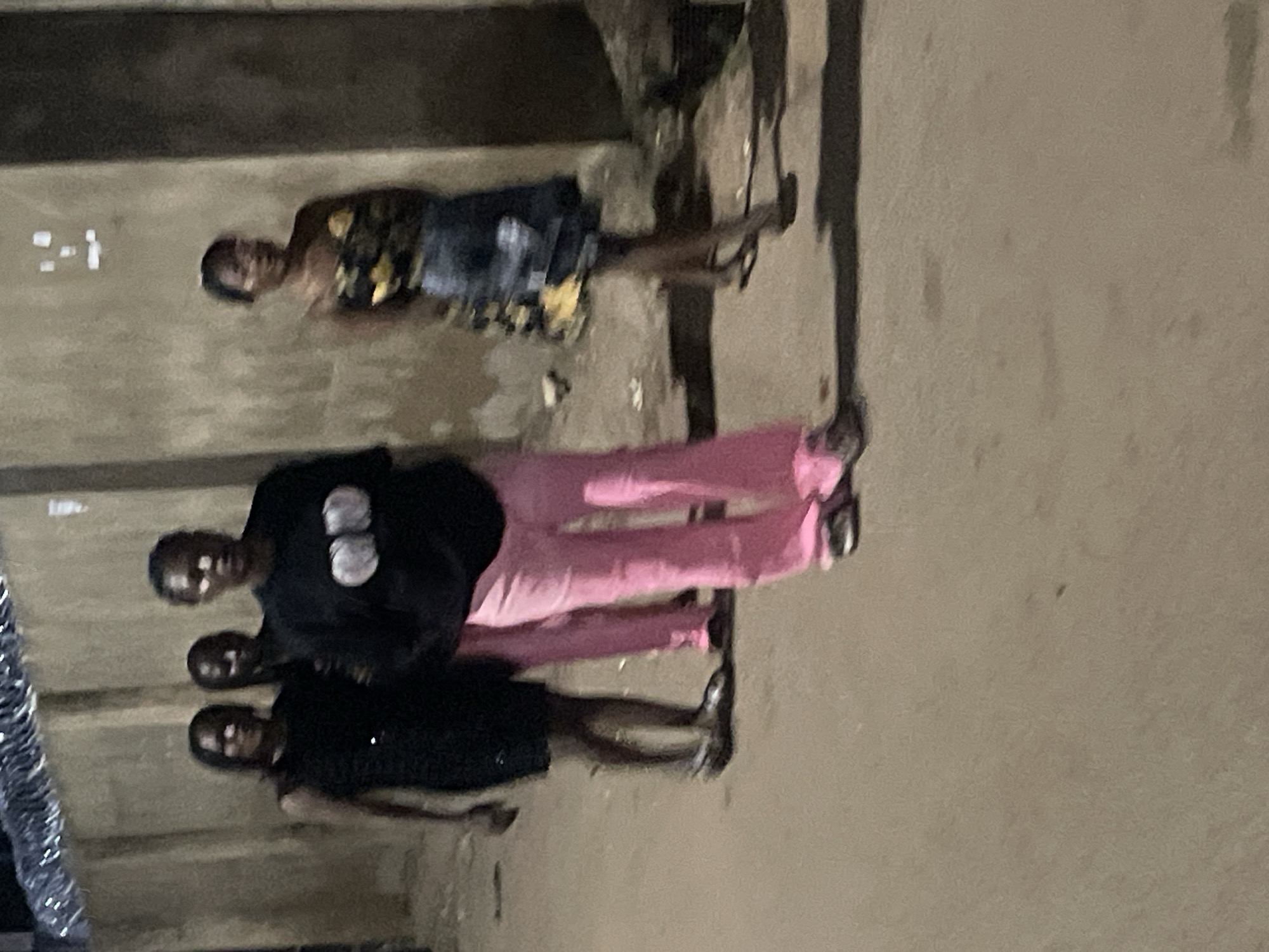 In the city of Jos, Nigeria, four individuals are captured in a nighttime photograph walking on a paved street. The individuals, appearing to be young women, are dressed in casual attire. The woman in the foreground on the left wears a black dress, while the woman next to her is in a black t-shirt with a graphic design and bright pink pants. To the right, another woman is seen in a floral pattern top and a denim skirt, and the fourth individual is partially obscured behind the others. The environment suggests an urban setting, with a textured wall in the background and what appears to be barbed wire or fencing at the top of the wall. The lighting is dim, indicating it is likely nighttime, and the focus is soft, giving the image a slightly blurred appearance.