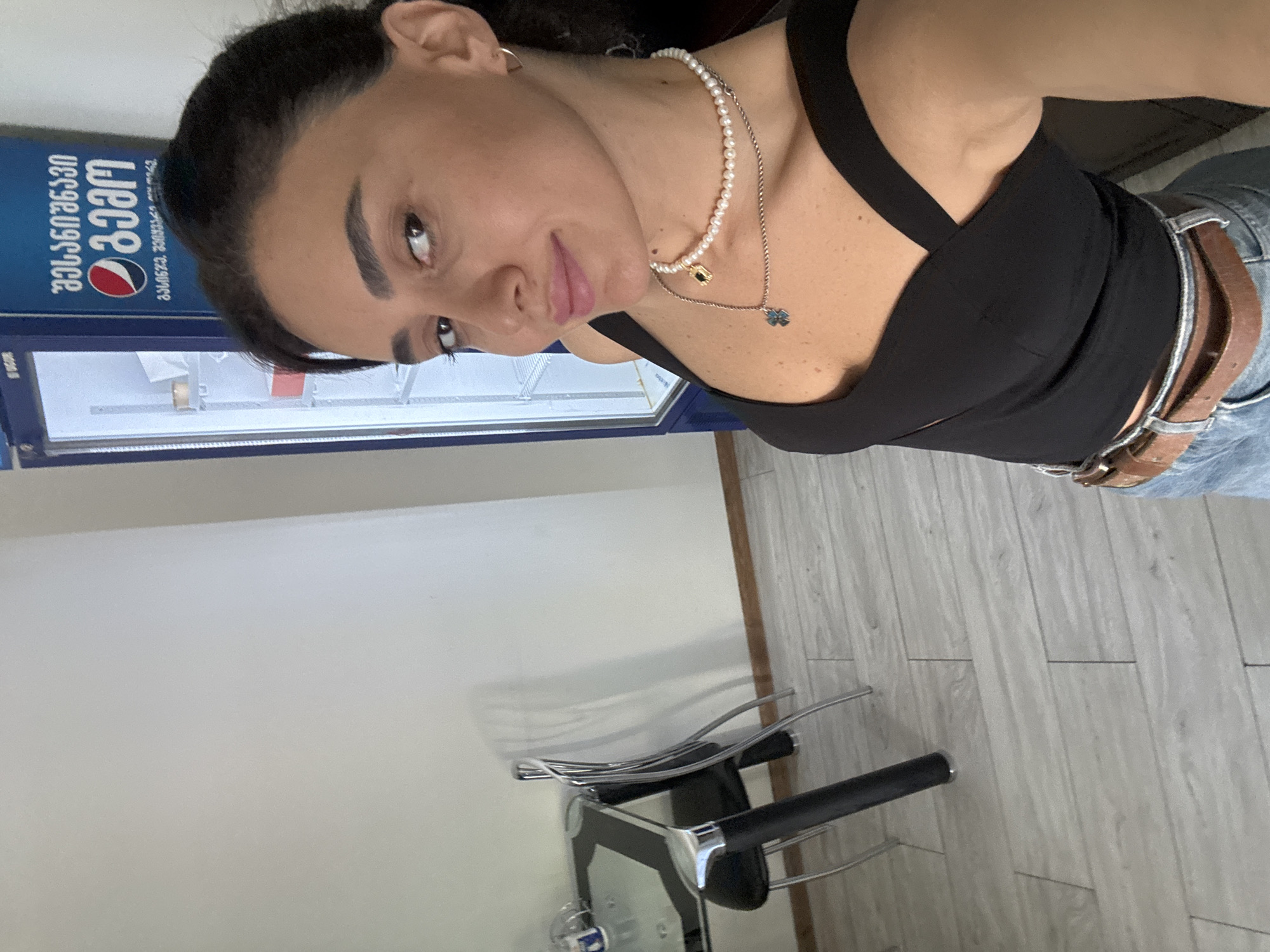 In a room in Tbilisi, Georgia, a woman takes a selfie in front of an open Pepsi cooler. She is wearing a black top and blue jeans, with a pearl necklace and a pendant. A glass-top table and chairs are visible to her left, and the room has light-colored walls and wood-like flooring.