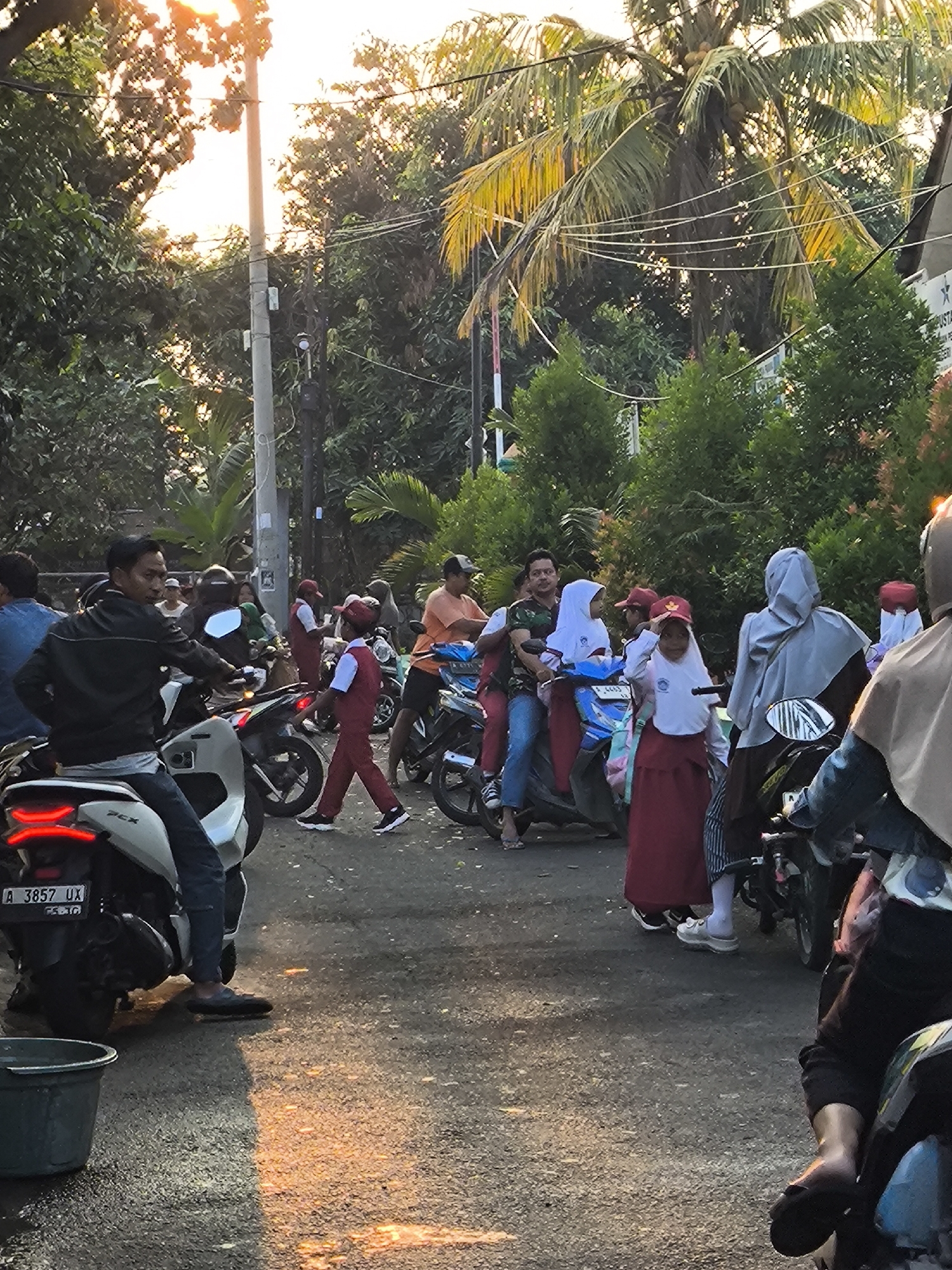 In Cilegon, Indonesia, at street level with a backdrop of lush green trees and the warm glow of sunset filtering through, a diverse group of individuals is gathered. The scene appears to be a busy intersection or roadside, with several motorcycles parked and people interacting. In the foreground, a man wearing a black leather jacket and jeans is seated on a white motorcycle. To his left, a dark grey bucket sits on the ground.
Several individuals, including children dressed in school uniforms, are present. One child, a girl in a red skirt and white top with a red cap, is standing and appears to be saluting or gesturing. Other children are on or near motorcycles, some accompanied by adults. One woman, wearing a light grey hijab and a dark garment, is standing next to a motorcycle. Another woman, wearing a beige hijab and dark clothing, is seated on a motorcycle in the far right foreground, her arm resting on the handlebars. A man in an orange shirt is standing near a motorcycle further back. The environment is characterized by a paved road reflecting the sunset, overhead power lines, and the presence of tropical foliage, including palm trees with coconuts. The overall impression is a snapshot of everyday life in a community, possibly during the end of the school day or a commute.
