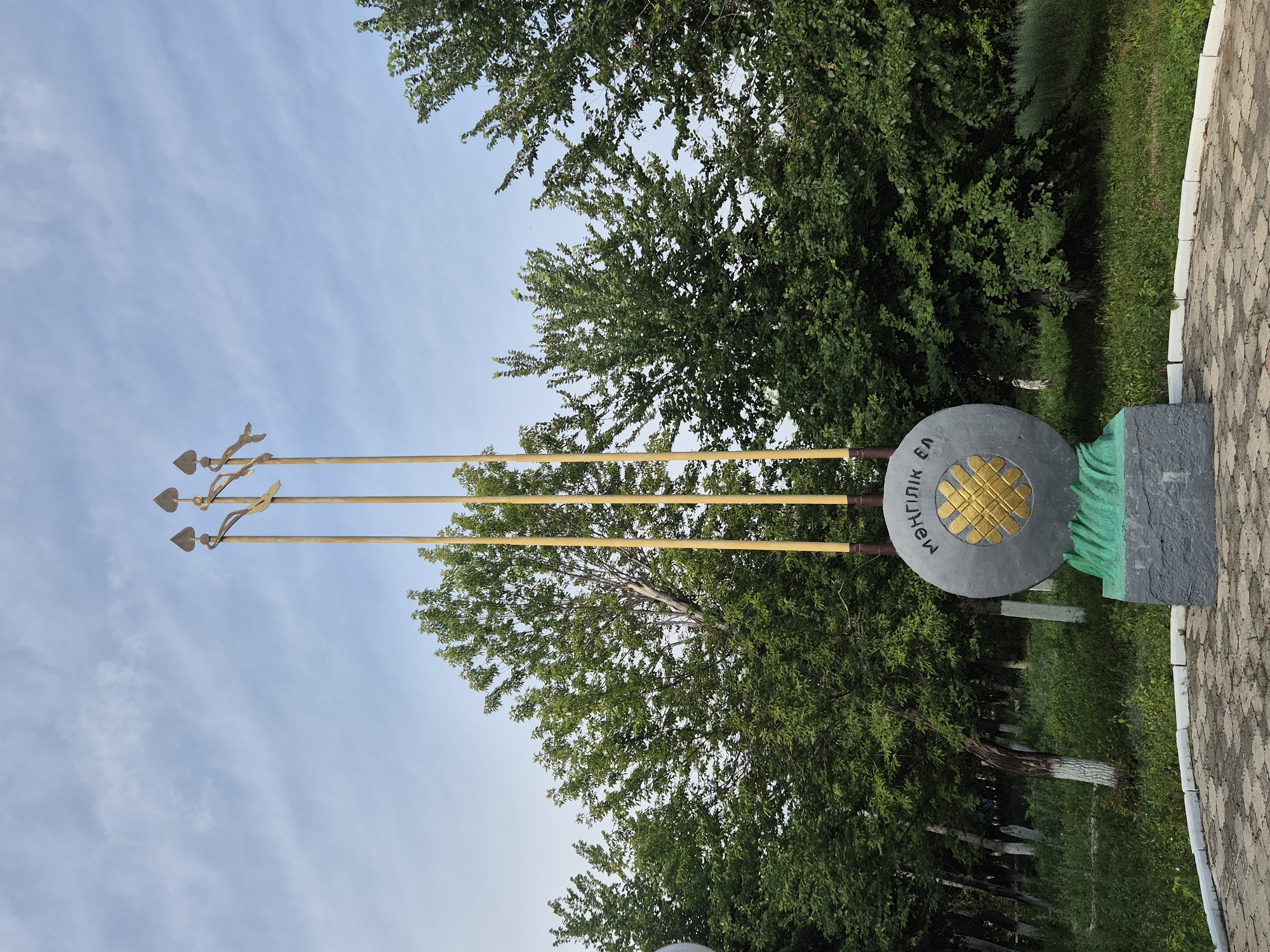 The image shows a monument in the city of Novoishimskoye, Kazakhstan. The monument consists of three tall, slender yellow poles topped with what appear to be spearheads. Between the poles, and closer to the ground, is a large, circular gray stone with a gold symbol in the center, resembling a geometric flower or star. The stone has Cyrillic lettering inscribed around its circumference. The monument is situated on a paved area, with lush green grass and trees surrounding it. The sky above is a pale blue with scattered white clouds.