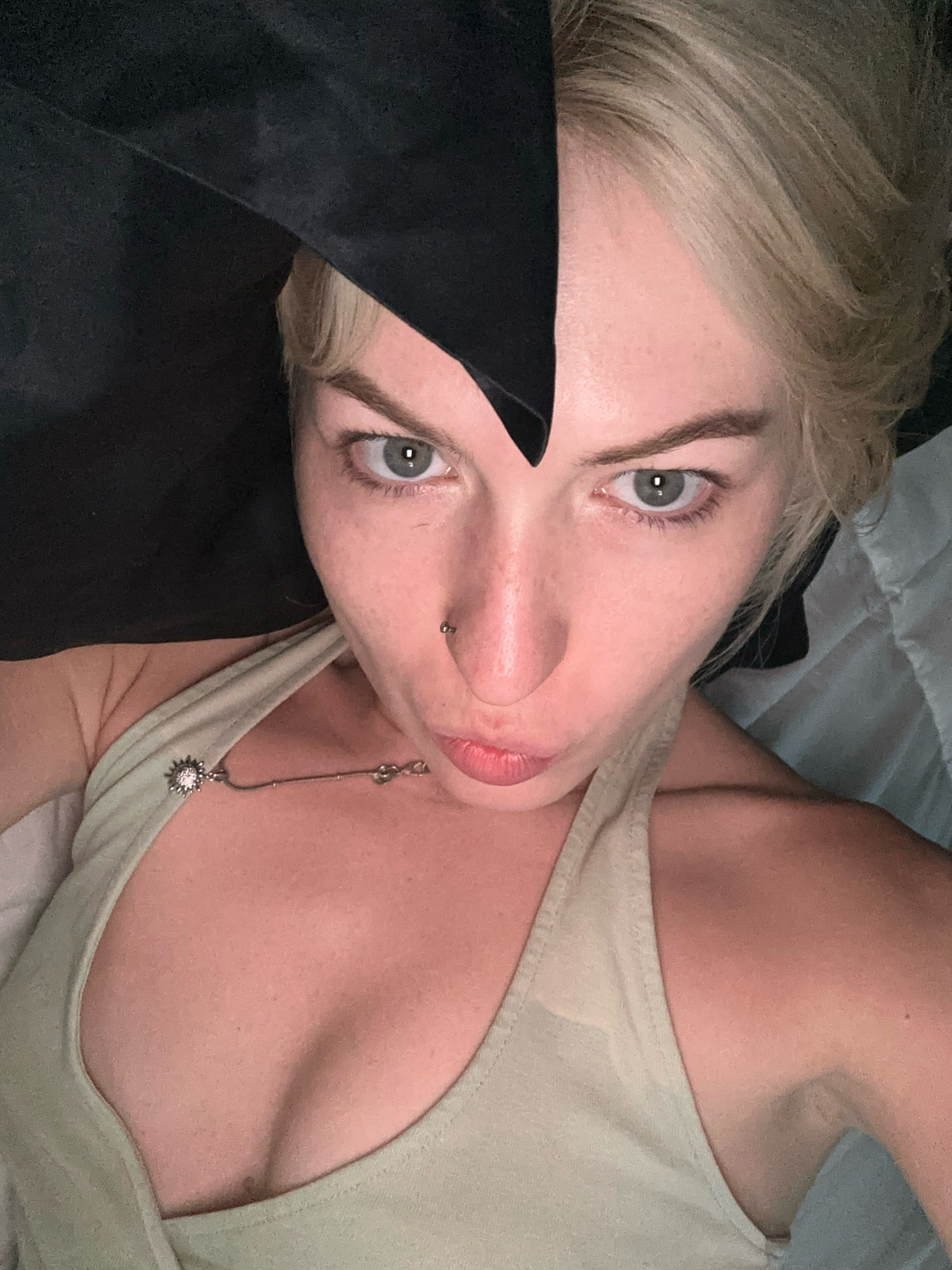A close-up, overhead shot shows a woman with blonde hair and fair skin. She is wearing a light-colored tank top, and a black fabric, possibly a hood or head covering, is draped over her head and shoulder. Her left eye is visible, looking at the camera. A nose ring is visible on her left nostril. The background appears to be a bed with white linens and a dark surface. The image is taken indoors, and there is no visible indication of the location in Tbilisi, Georgia.