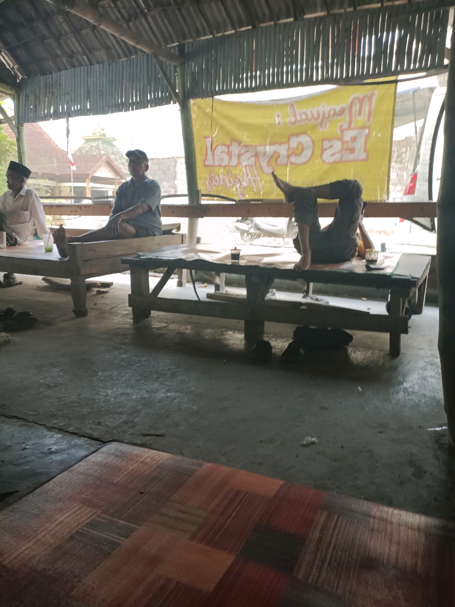 In Rancang Kencono, Indonesia, a dimly lit open-air shelter with a corrugated metal roof and wooden supports houses a scene of casual repose. Two men are seated on low wooden platforms. The man on the left wears a white long-sleeved shirt and a dark peci, appearing to be engaged in conversation. The man in the middle wears a grey t-shirt and a dark baseball cap. To his right, a third individual, dressed in dark pants, is sprawled on a wooden table with their legs bent and extended upwards, resting their head on their arms. A yellow banner with Indonesian text, likely an advertisement, hangs behind them. On the table in front of the sprawled individual, a small glass with a red liquid and a straw is visible, along with other indistinct objects. The floor appears to be a concrete surface, with a woven mat in the foreground. The overall atmosphere suggests a relaxed, possibly informal gathering place.