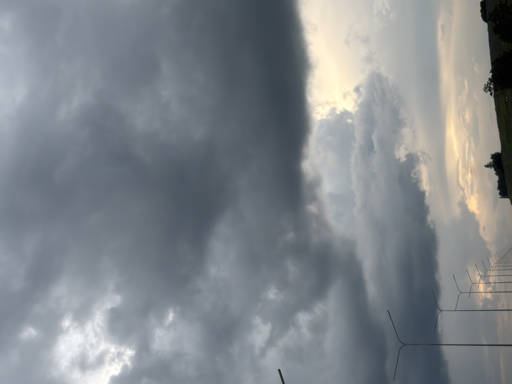 The image shows a street scene in Кояново, Russia, featuring a row of streetlights stretching into the distance. The sky is filled with dark, heavy storm clouds, suggesting an impending downpour. The lighting is dim, with a sliver of brighter sky visible near the horizon, possibly indicating the sun attempting to break through. The foreground is dominated by the stark silhouettes of the streetlights, emphasizing their repetitive nature along the road. A grassy embankment with a few scattered trees can be seen in the far distance, adding a natural element to the otherwise urban infrastructure. There are no people visible in the image. The overall atmosphere is one of a cloudy, possibly humid, day, characteristic of a storm's approach.