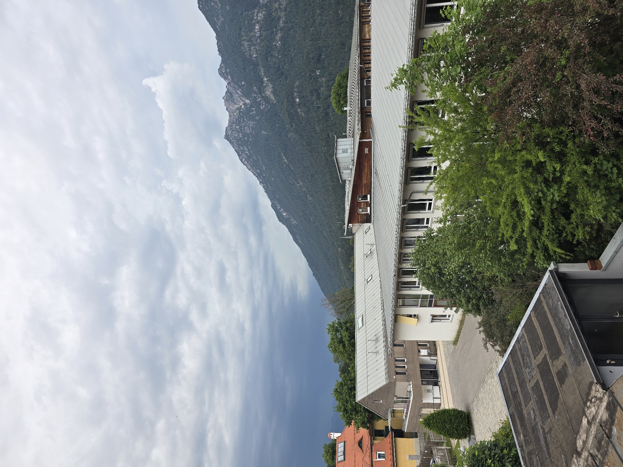The image captures an aerial view of buildings and a mountain range under a cloudy sky in Garmisch-Partenkirchen, Germany. In the foreground, several roofs are visible, one with a dark, weathered surface. Beyond these, a long, modern building with a silver roof and numerous windows stretches across the frame. To the left of this building, a smaller structure with a reddish-orange roof and yellow siding is partially visible. Behind the buildings, lush green trees form a dense line. The backdrop is dominated by a majestic mountain, its slopes covered in dark green forest. The sky above is overcast with dramatic dark clouds gathering on the left, suggesting approaching rain, contrasting with the lighter, diffused clouds elsewhere. No people or animals are discernible in the scene.