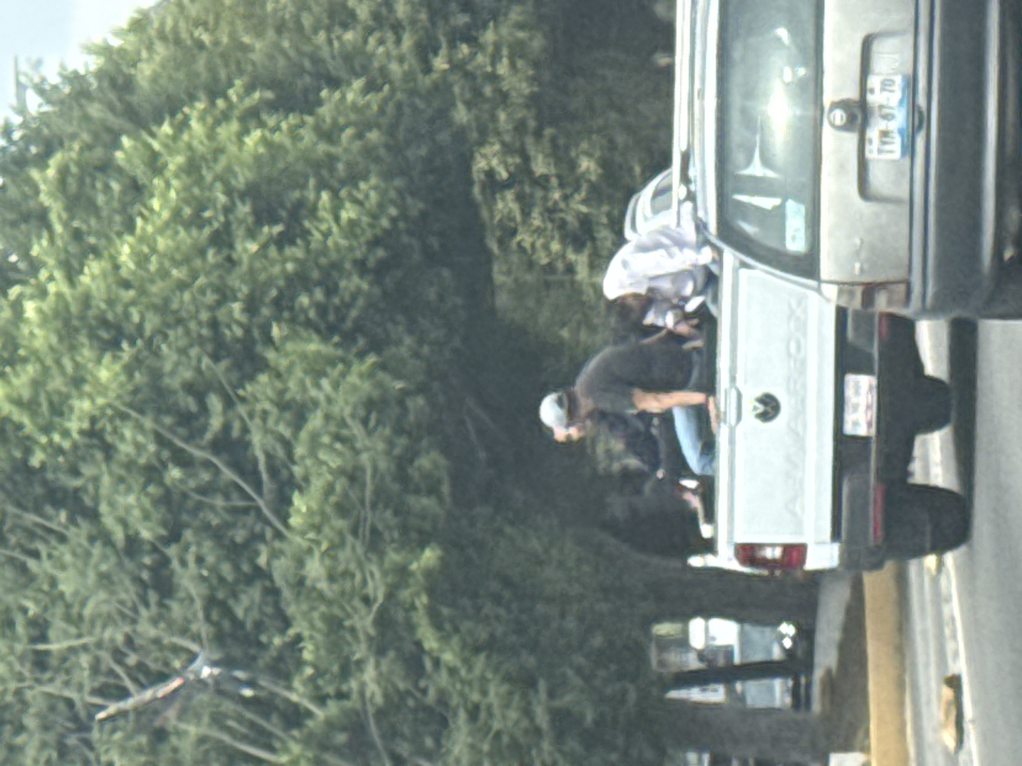The image depicts two people standing in the bed of a parked, white Volkswagen Amarok pickup truck. One person is wearing a dark shirt, jeans, and a baseball cap, while the other is wearing a white shirt. The license plate of the vehicle reads TTM-67-70. The truck is parked on a street next to a sidewalk, with a large green tree in the background. Additional vehicles are visible further down the street. The location is Puebla, Mexico.