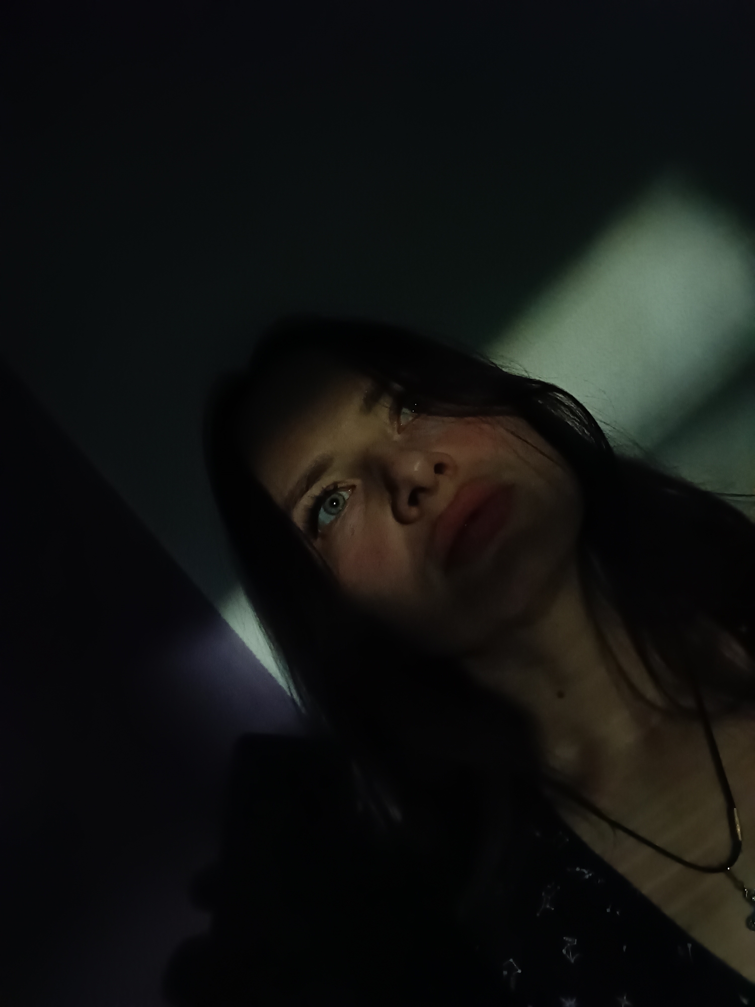 A low-angle, close-up portrait features a young woman with dark hair against a dark, shadowy background. The scene is dominated by deep shadows, with a strong, diagonal beam of light illuminating the upper left portion of the frame and parts of the woman's face.
The woman is positioned in the lower right half of the image, looking upwards and slightly to the viewer's left. Her left eye, a striking blue or green, is clearly visible and catches the light, while her right eye and much of her face are enveloped in shadow. Her lips are slightly parted, and her expression appears pensive or melancholic. Her hair is dark and falls around her face and shoulders. She wears a dark garment, possibly a top or dress, with faint, subtle patterns visible in the dimly lit areas near her chest. A thin, dark necklace with a small, metallic pendant is visible around her neck.
The background is an indistinct dark wall or interior surface, with the diagonal light creating a prominent feature. The overall mood is somber and introspective, emphasized by the dramatic lighting and shadow play. No specific activity is depicted other than posing for the photograph. There is no visible text. The setting appears to be indoors in Rusalevka, Russia, with the distinct lighting suggesting artificial illumination.