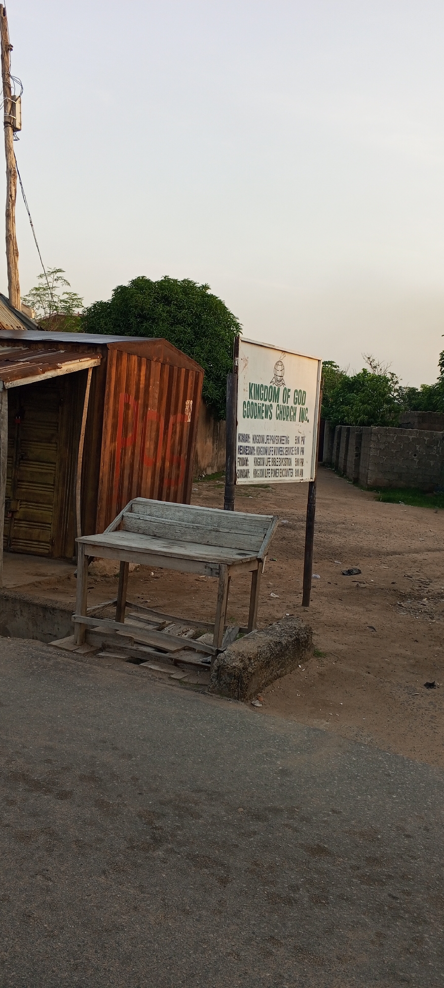 The image shows a street scene in Jalingo, Nigeria. In the foreground, a cracked asphalt road is visible. To the right of the road, a dirt path leads away from the viewer, bordered by a low wall constructed of grey concrete blocks. On the left side of the image, a dilapidated wooden bench sits in front of a rusty corrugated metal structure. Further down the dirt path, a white sign with black lettering displays information about "KINGDOM OF GOD GOODNEWS CHURCH INC." and lists various meetings and their times. Behind the church sign, lush green trees and bushes create a backdrop. A wooden utility pole with wires is positioned to the far left of the frame. The sky is a pale, hazy blue, suggesting it may be early morning or late afternoon. The overall scene depicts a simple, somewhat weathered urban environment.