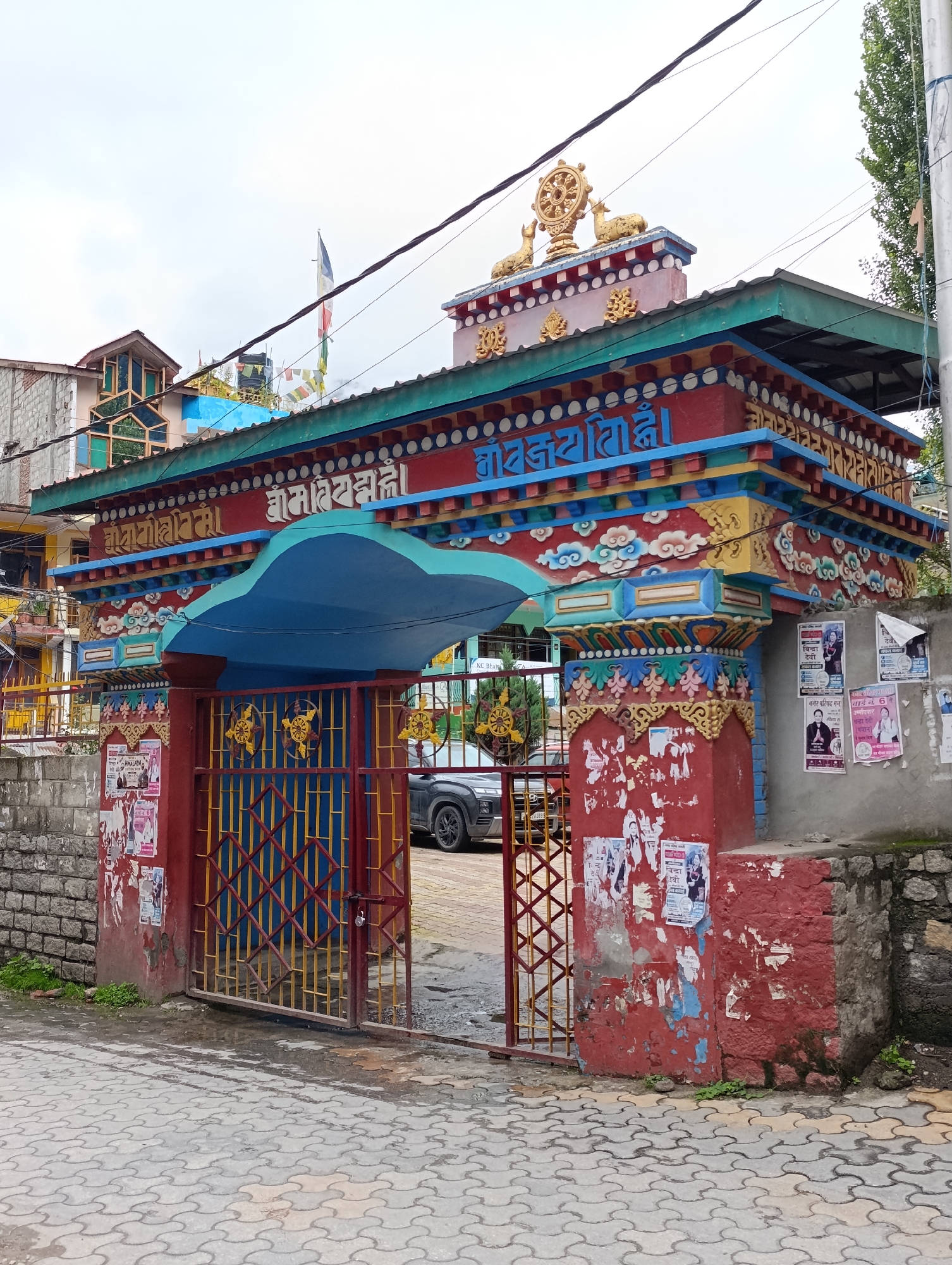 The image shows the entrance to the Tashi Lhunpo Monastery in Manali, India. The entrance is a brightly painted gateway with colorful decorations and Buddhist symbols. Prayer flags are visible fluttering above the gateway. A red and yellow metal gate is partially open, revealing a glimpse of the monastery's interior, including a parked car. The surrounding area is paved with interlocking stones, and there are some posters plastered on a red-painted wall to the right of the entrance. The sky is overcast.
