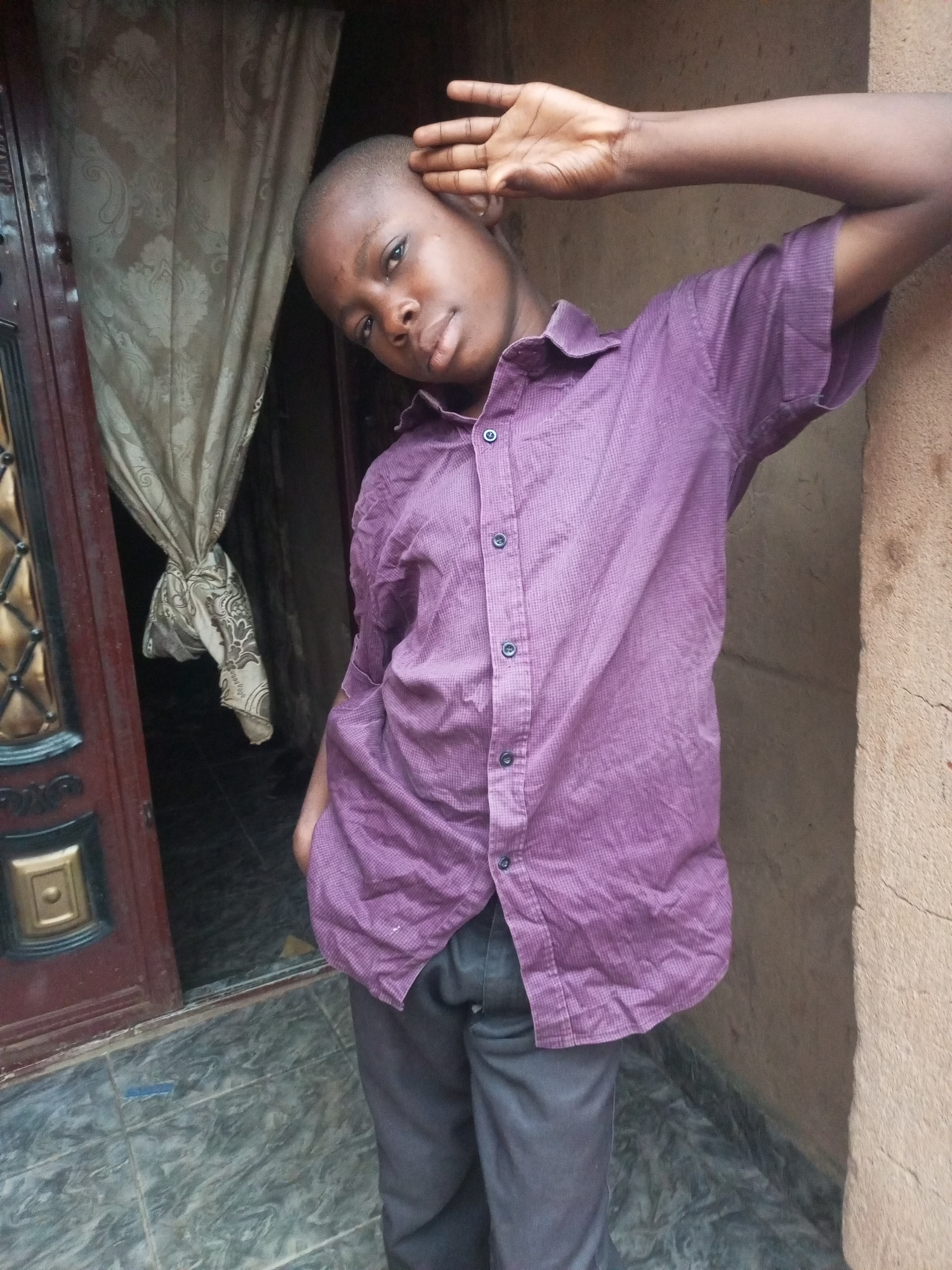 A young Black boy with short hair stands in front of a textured wall, leaning to his right. He is wearing a purple, short-sleeved, collared shirt with small dark buttons. His right arm is raised, with his hand resting on his forehead or temple. His left hand is in his pocket. The background includes a brown textured wall on the right, and on the left, a dark doorway with a partially opened, dark red door. The door has decorative gold and black embellishments and a tan, patterned curtain hanging inside. The ground in front of the door and wall is tiled with a grey and white marbled pattern. The scene appears to be outdoors, possibly in an urban environment like Tempure, Nigeria, given the architectural style.