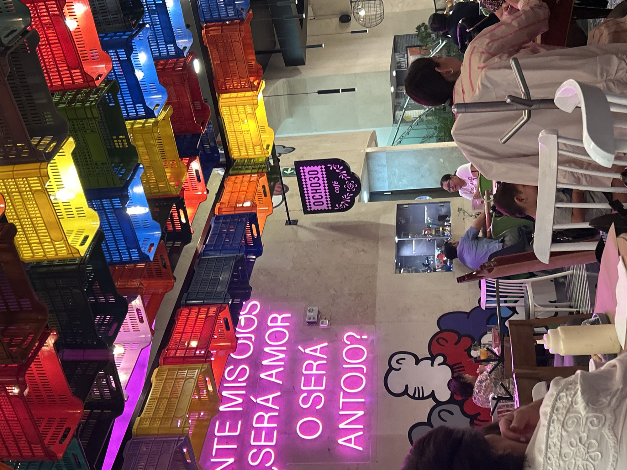 The image captures a scene at the Ocho30 Cafe in Puebla, Mexico. The interior decoration includes a colorful ceiling made from plastic crates and a neon sign that reads "ANTE MIS OJOS SERA AMOR O SERA ANTOJO?". Patrons sit at tables, appearing to be dining. The environment seems casual and vibrant.