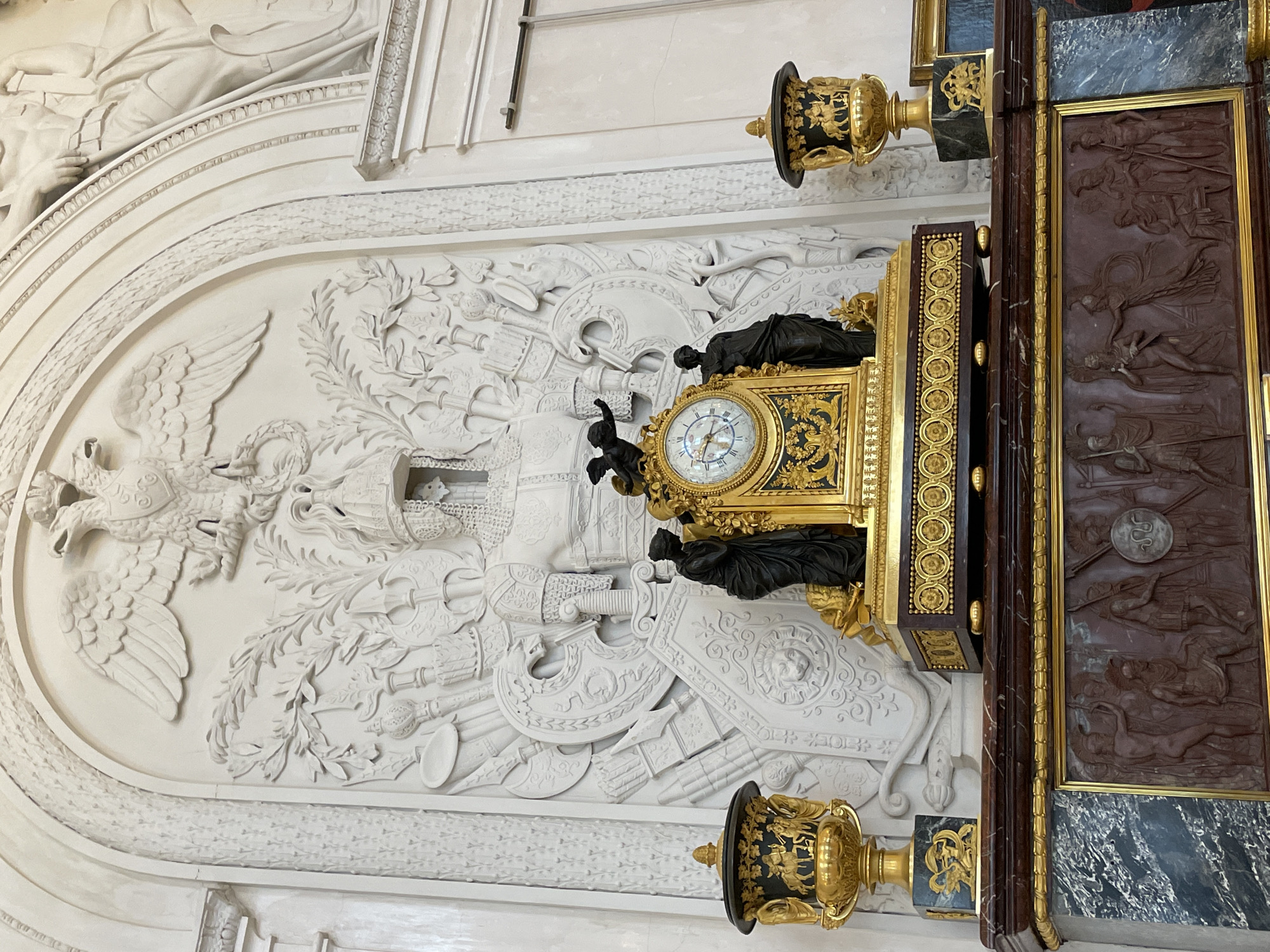 The image shows a wall in an unknown city. On the left, a white wall features an oval-shaped relief of an eagle and a collection of swords, shields, and armor. To the right, a large decorative clock is positioned, with black statues flanking the clock face and ornate golden embellishments around it. Flanking the clock are two gold and black urns or lamps. To the far right, a dark brown paneled wall shows carvings of people in motion. The scene appears to be indoors, potentially in a museum or historical building.