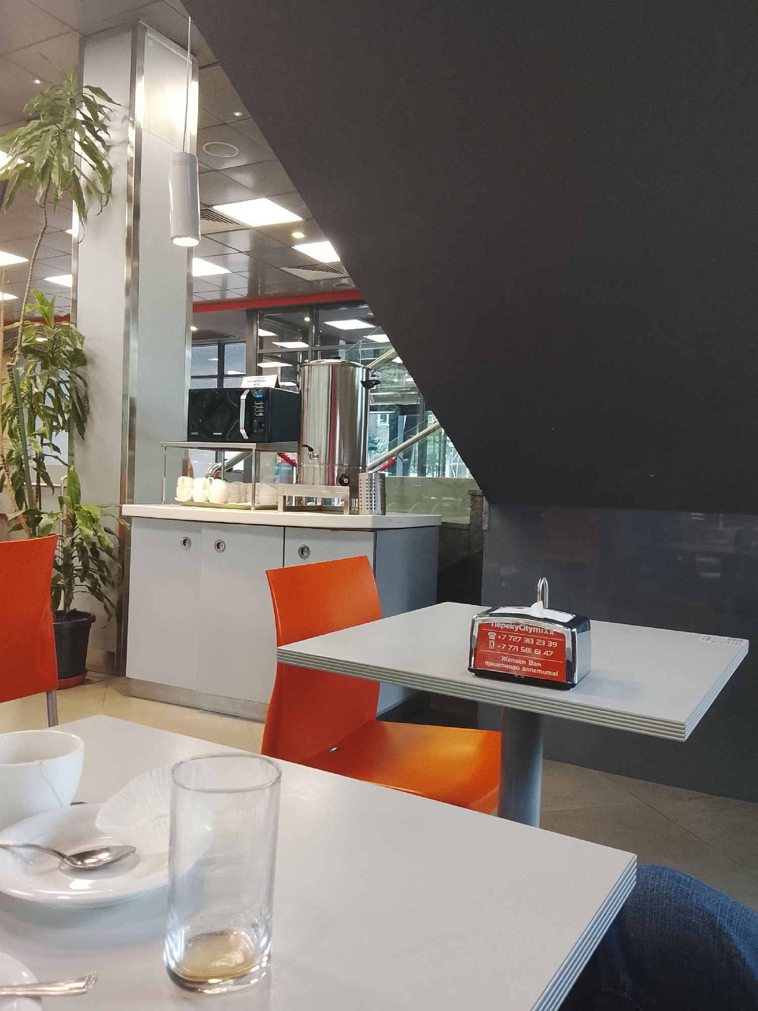 The image shows the interior of a cafe or dining area. In the foreground, there is a white table with a white cup, a saucer with a spoon, and an empty drinking glass. Further back, there is a counter area with a microwave, a large metal urn, and several white cups. Orange chairs are visible on either side of the table. In the background, a large black diagonal structure dominates the upper portion of the image, with ceiling lights visible above. The overall environment suggests a modern, possibly casual dining establishment. No specific location in Almaty, Kazakhstan, can be determined from the image alone, as it lacks any identifiable landmarks or signage indicating a precise location within the city.