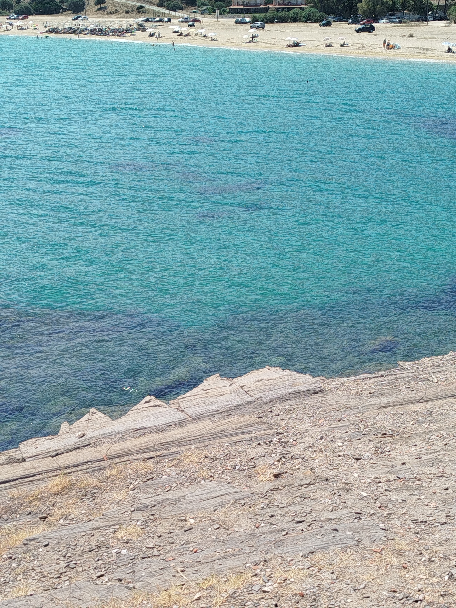 The image shows a view of a coastal area in the Sithonia Municipal Unit, Greece. The foreground features a rocky, sloping terrain covered with small stones and sparse dry vegetation. The midground is dominated by clear, turquoise-colored water with gentle waves, revealing the seabed beneath in shallower areas. In the background, a sandy beach is visible, lined with numerous beach umbrellas and sun loungers. Several people are scattered across the beach, some sitting under umbrellas and others standing near the water. A row of parked cars is situated behind the beach, adjacent to a tree-lined area. The overall scene depicts a tranquil summer day at a beach resort.
