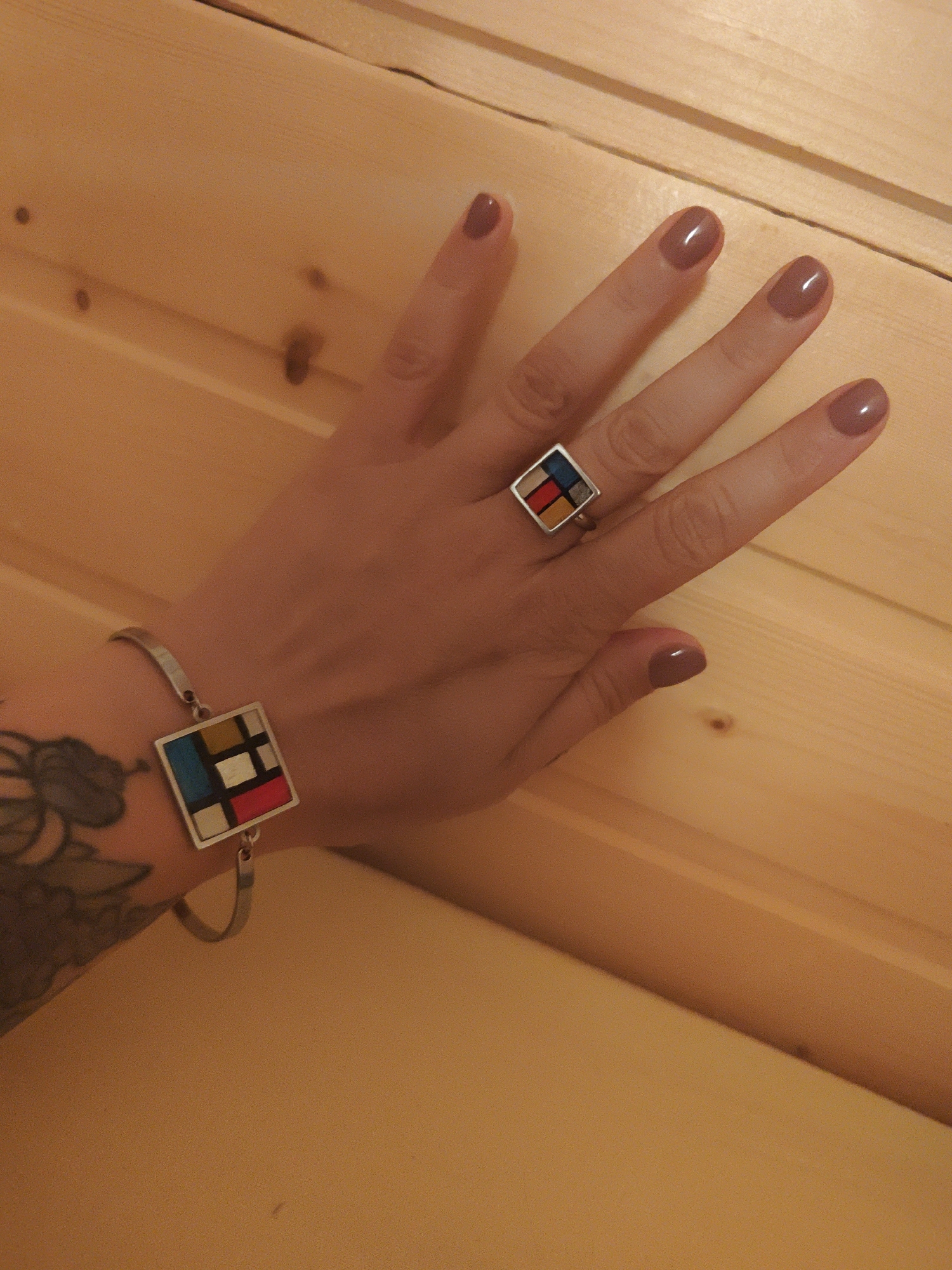The image displays a person's left hand and lower forearm resting on a light-colored wooden surface, likely a table or wall composed of planks. The hand is adorned with two pieces of jewelry, both featuring a square design inspired by Piet Mondrian's artwork. A silver-toned ring with a square, segmented face showcasing blocks of red, blue, yellow, white, and a dark color (possibly black or dark grey) is worn on the middle finger. A matching silver-toned bracelet with a similar square, multi-colored segment design is on the wrist. The fingernails are neatly manicured and painted a glossy, muted purplish-brown or taupe color. A dark, intricate tattoo is partially visible on the inner side of the forearm. The wooden surface beneath the hand exhibits a natural grain and several dark knots, suggesting a warm and rustic indoor setting. The lighting is soft and warm, typical of indoor artificial light. No specific activity is occurring; the hand is simply posed, showcasing the jewelry and nail polish. No text is visible in the image.