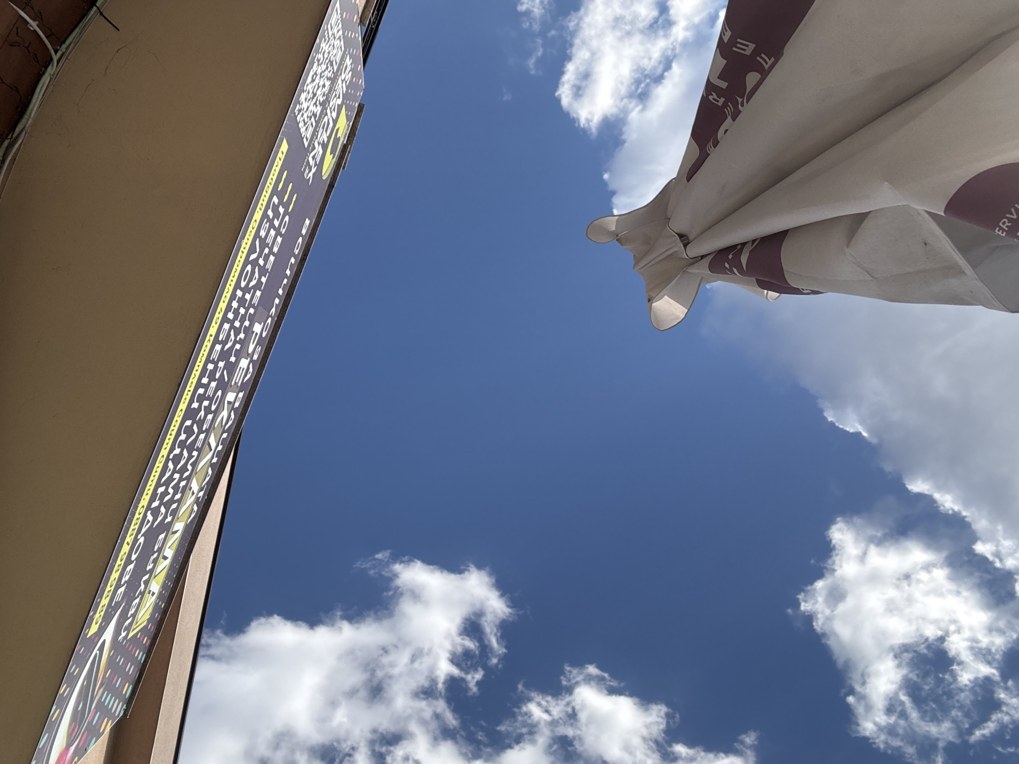 The image shows a low-angle view of the sky in Plovdiv, Bulgaria, partially obscured by clouds and two architectural elements. A tan building with a vertical sign is visible on the left, and a white umbrella with a burgundy pattern is seen on the right. The sky is predominantly blue, with scattered white clouds. The sign is mostly unreadable due to the angle and depth of field.