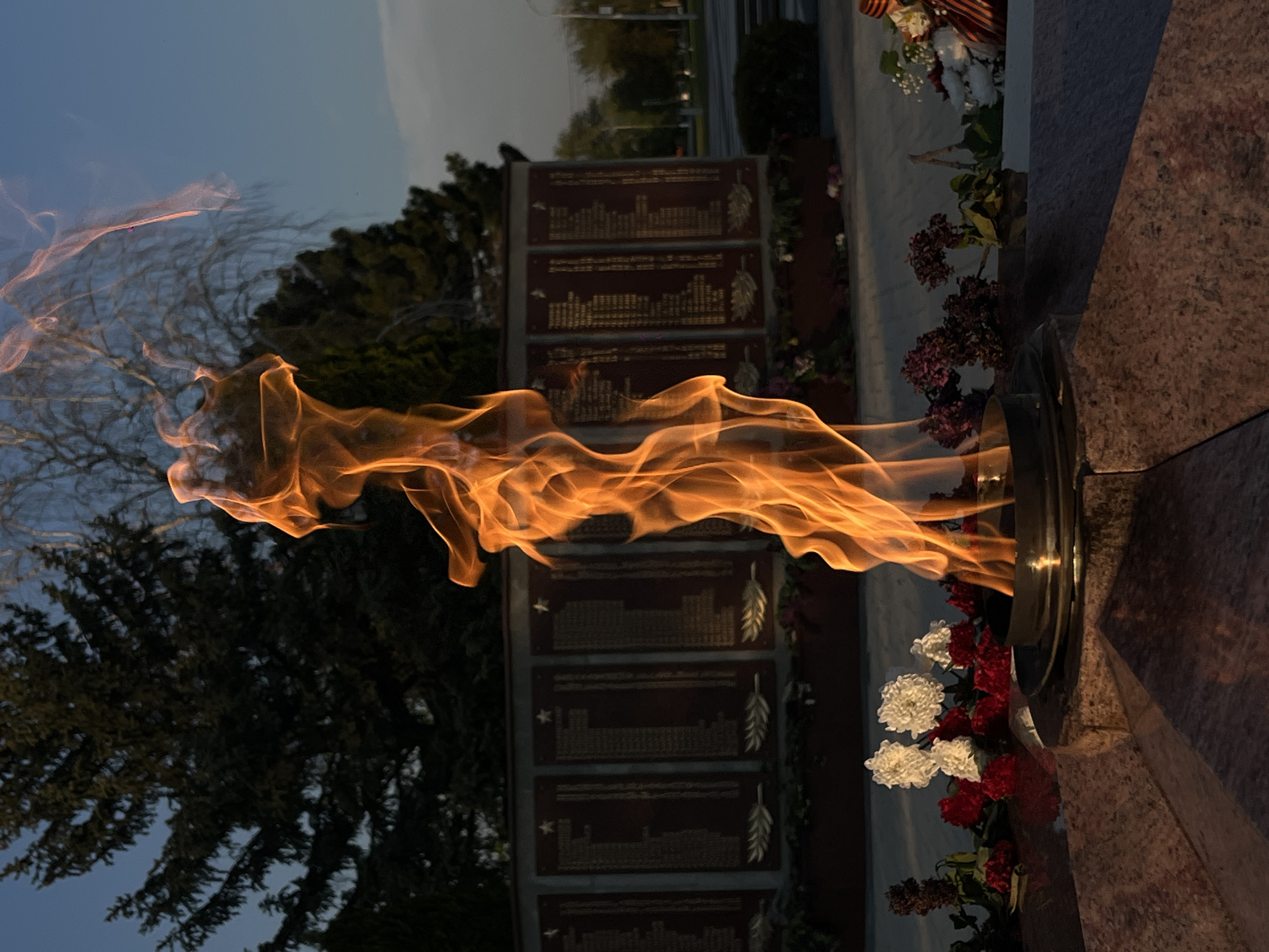 The image shows an eternal flame burning horizontally. The flame extends to the left, with an orange and yellow hue, set against a darkening sky. Behind the flame, on the left, a tree is visible with dark green foliage. To the right of the flame are memorial plaques with inscribed text and decorative leaf designs. Flowers are placed around the flame’s base, including white and red blooms. The flame emerges from a dark metal structure, which is part of a stone monument made of a light-brown granite. The monument and flame are located outdoors, likely in Novopokrovskaya, Russia. The scene seems to be at dusk.