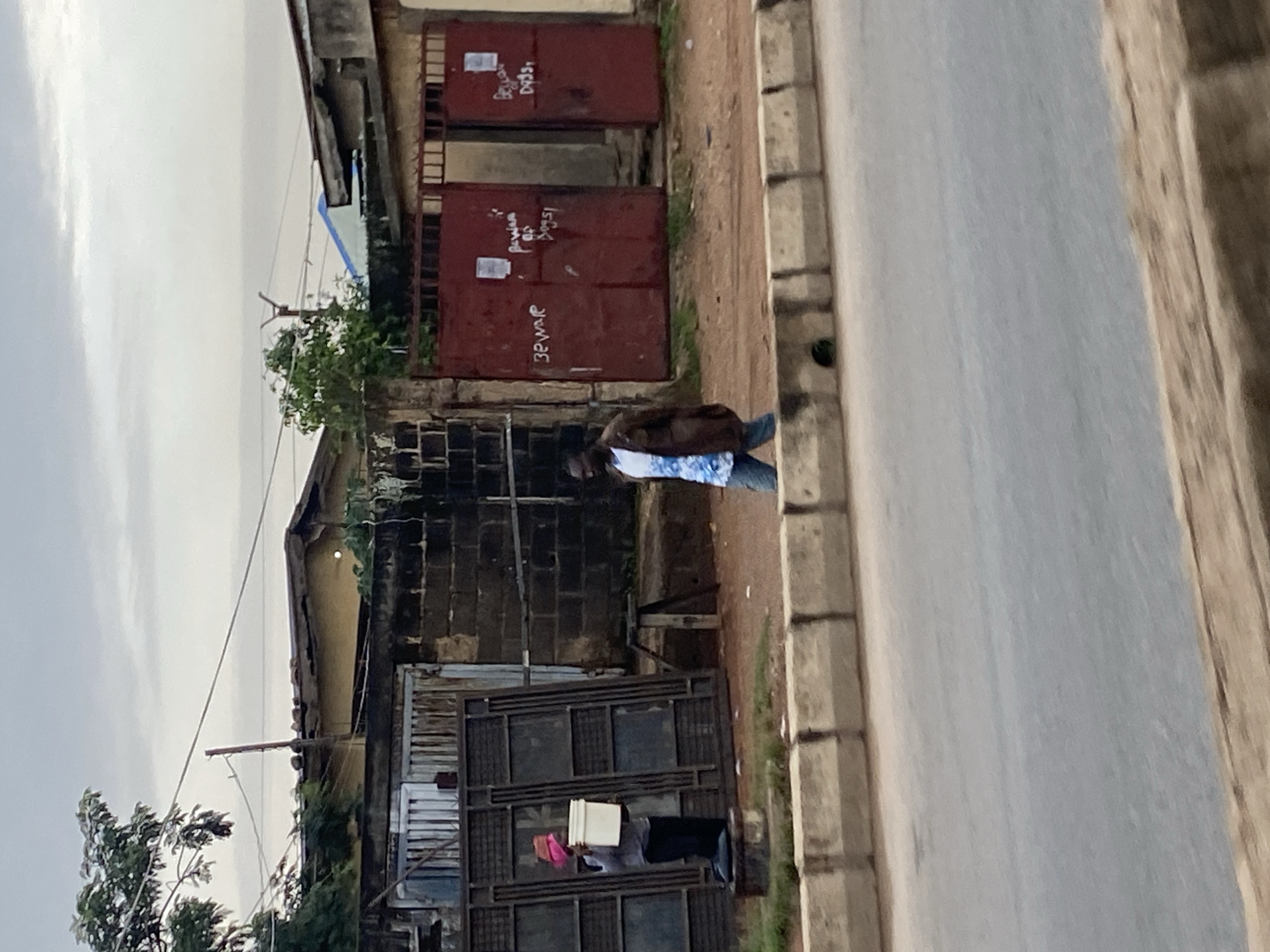 The image depicts a street scene in Jos, Nigeria, under an overcast sky. In the foreground, a paved road is bordered by a concrete median. Beyond the median, a sidewalk area is visible, with a person kneeling on a small concrete block, wearing a pink head covering and holding a white bucket. They are positioned in front of a tall, ornate metal gate. To the right, another individual walks away from the camera, wearing a blue and white patterned shirt under a brown jacket and dark pants. Behind these individuals, a wall constructed of dark bricks forms the boundary of a property. This wall is interrupted by two large, red metal gates, one of which is partially open. Both gates have white notices or signs affixed to them, and writing appears to be spray-painted on their surfaces, with words like "BEWARE" and "DOGS" partially legible. Structures with pitched roofs are visible behind the gates. Trees with dense foliage are present on the left side of the image, and utility poles with wires are also visible against the sky. The overall environment suggests a commercial or residential area in an urban setting.