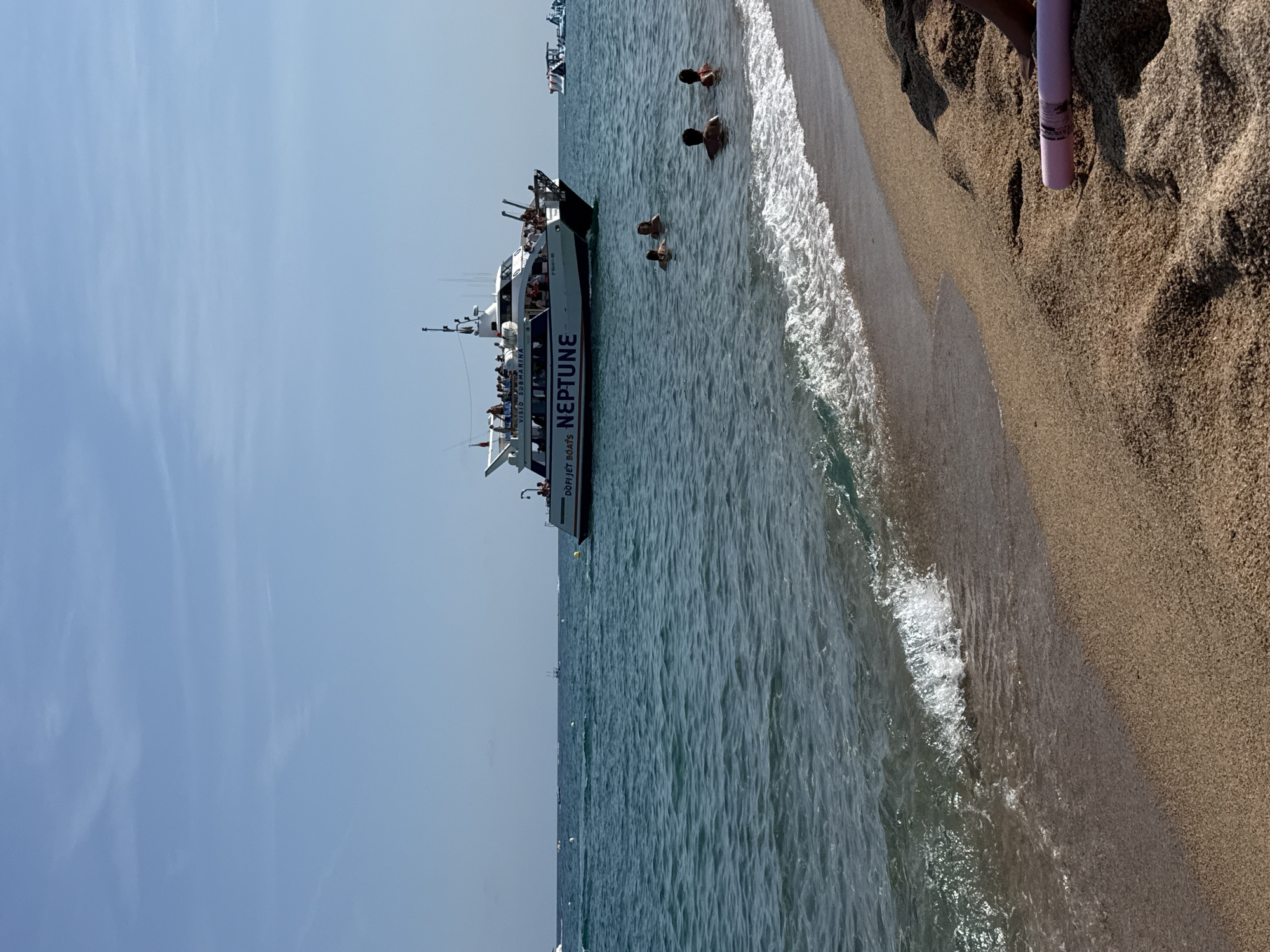 The image captures a sunny day at a beach in Blanes, Spain. In the foreground, a sandy beach slopes gently towards the turquoise water. Small waves are breaking on the shore. In the mid-ground, several people are swimming in the sea, with their heads and shoulders visible above the water. Further out, a large white boat named "NEPTUNE" is cruising, with many passengers visible on its decks. The boat appears to be a tour boat, possibly a glass-bottomed boat, as indicated by the text "VISIO SUBMARINA" on its side. In the background, the horizon separates the sea from a clear blue sky with a few wispy clouds. Other smaller boats and watercraft are scattered across the water. The overall scene depicts a lively beach day with recreational activities taking place in and around the water.