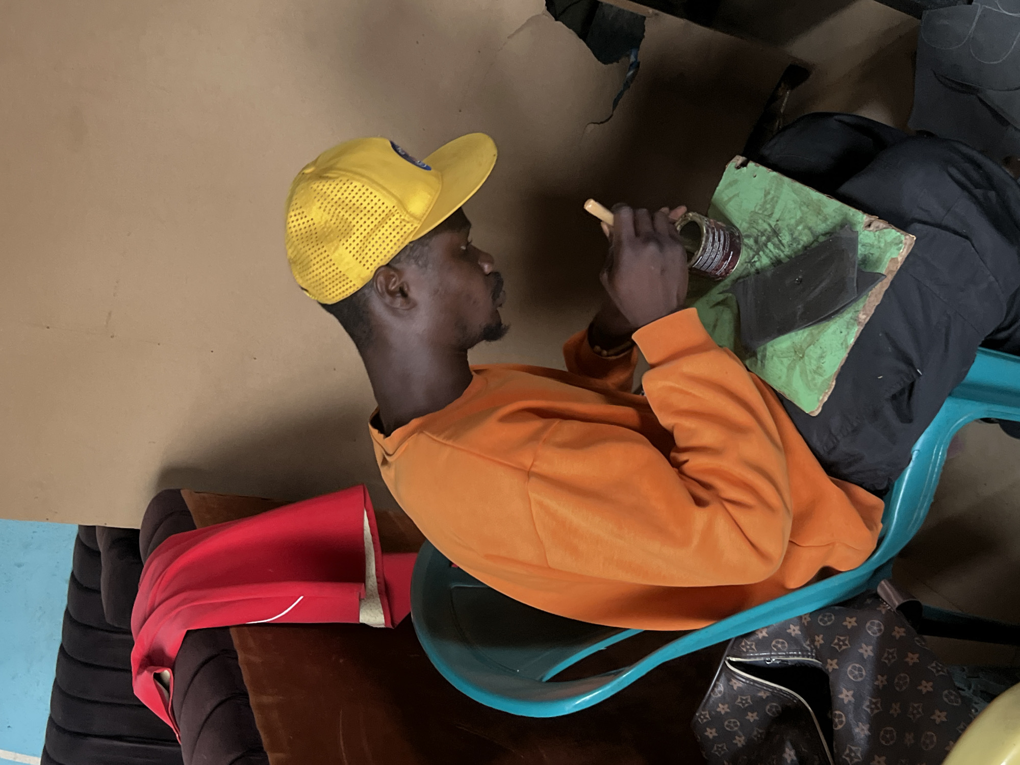 An indoor, portrait-oriented photograph captures a dark-skinned man seated on a teal-colored plastic chair, engaged in a craft or repair activity. The man is positioned facing to the right of the frame, looking downwards intently at the items he is working with. He wears a bright yellow mesh baseball cap and an orange long-sleeved shirt, and has a neatly trimmed beard and mustache.
In his hands, he holds a glass jar with a metallic lid, containing a dark, reddish substance, and a small wooden tool or stick. His left hand grips the jar, while his right hand holds the stick. Directly in front of him, resting on his lap or a work surface, is a worn green board or mat, on which a dark, irregularly shaped object, possibly made of leather or rubber, is placed. This arrangement suggests he might be involved in leatherwork, shoemaking, or a similar craft requiring adhesives or dyes.
The background features a plain, light brown wall or floor. To the left of the man, a red fabric item, possibly a blanket or folded garment, rests on a dark brown cushioned surface, which appears to be part of a couch or another piece of furniture. Below his chair, on the floor, a brown bag with a repeating pattern of stars and circles (resembling a designer logo) is partially visible.
The lighting is consistent, indicating an indoor setting, likely illuminated by artificial light. The man appears focused and calm while concentrating on his task. No discernible text is visible in the image. The scene portrays a moment of diligent craftsmanship in a modest indoor environment, possibly in Jos, Nigeria.
