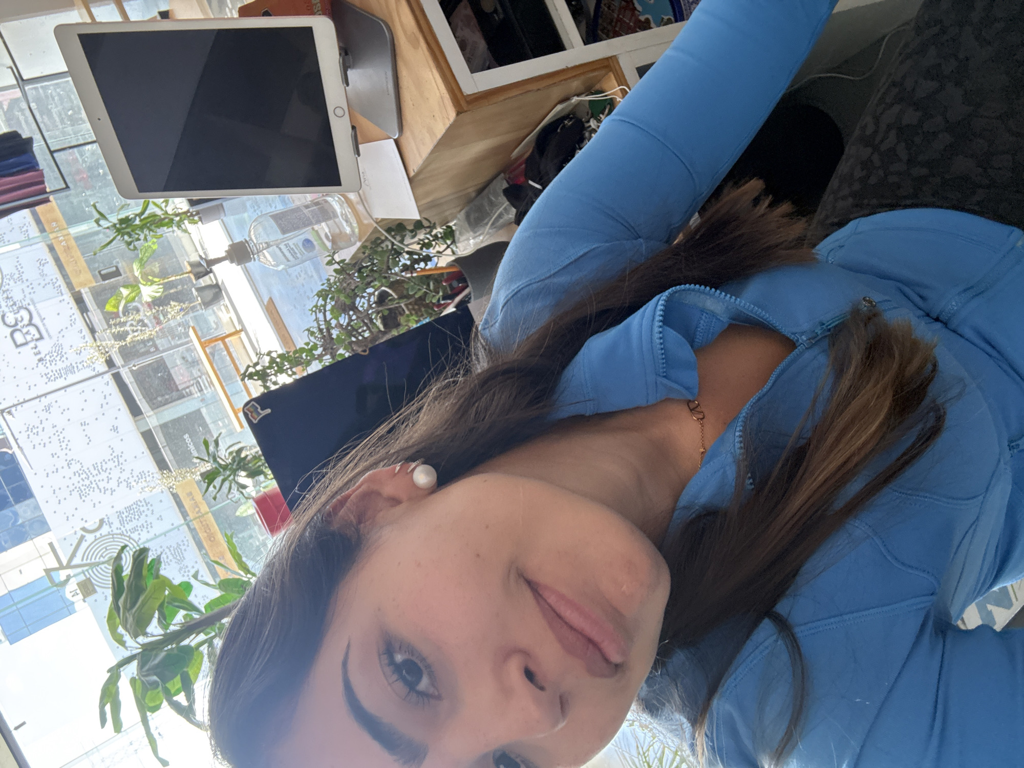 The image shows a selfie of a woman with brown hair, wearing a light blue zip-up jacket and a gold necklace. She has a pearl earring on her left ear. In the background, there is a window with a view of a city, plants, and a table with an iPad, hand sanitizer, and other items. The window view is likely Puebla, Mexico due to the distinctive architectural style.