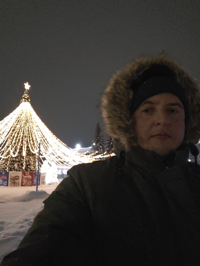 A man in a dark winter coat with a fur-lined hood stands outside at night. Behind him, a large Christmas tree is decorated with many strings of bright white lights and topped with a star. The ground is covered in snow. Other trees are visible in the background. The man looks toward the camera.