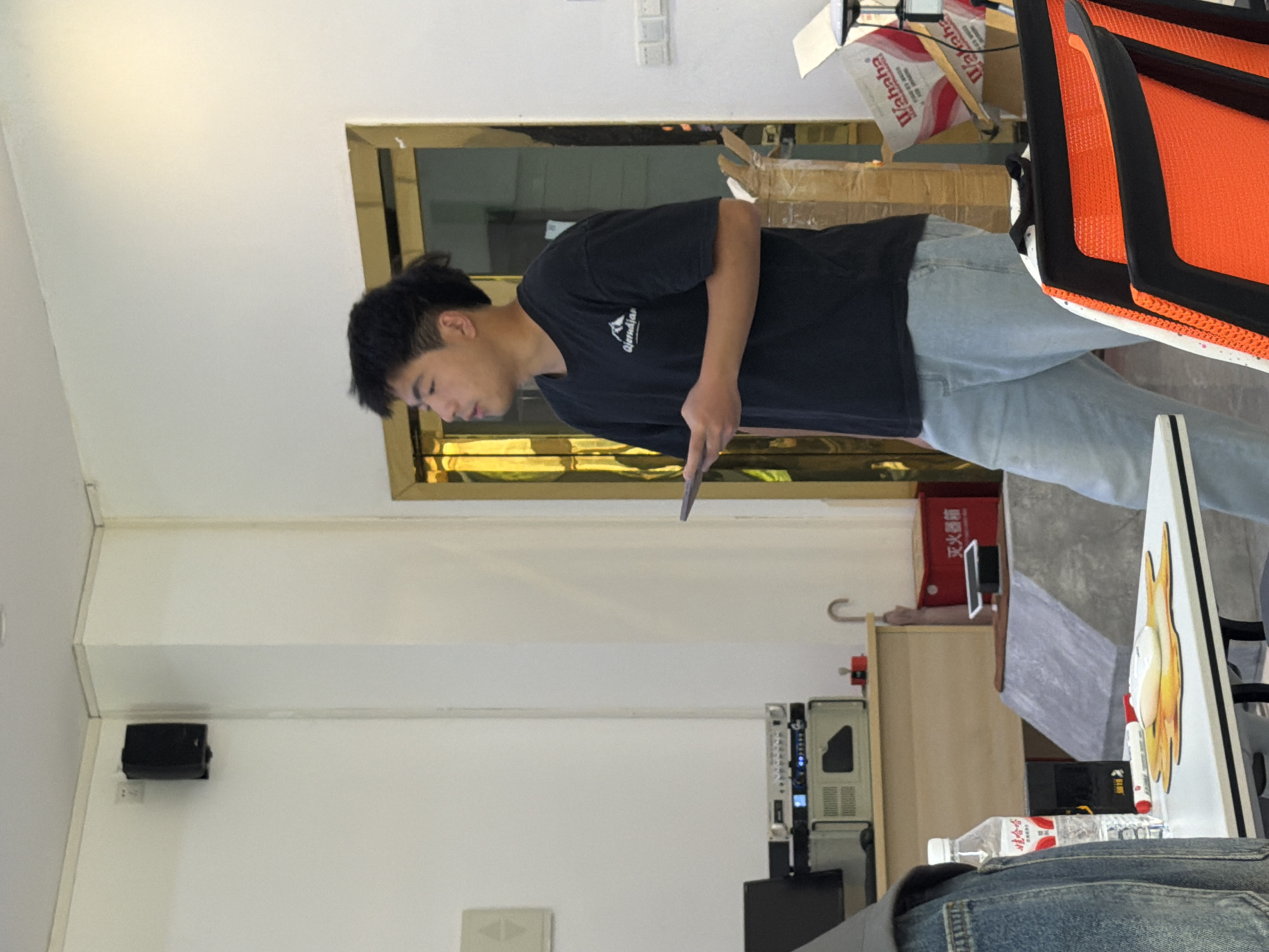 A young East Asian man wearing a black t-shirt and light blue jeans stands in a brightly lit room, holding a tablet in his right hand. He is looking to his right, with his mouth slightly open. The room appears to be an office or meeting space, with white walls and a wooden desk in the background. On the desk are various electronic devices, including a computer monitor and what appear to be audio equipment. To the right of the man, a stack of cardboard boxes is visible. The overall scene suggests an informal gathering or presentation. The image was captured in Jiangjin, China.