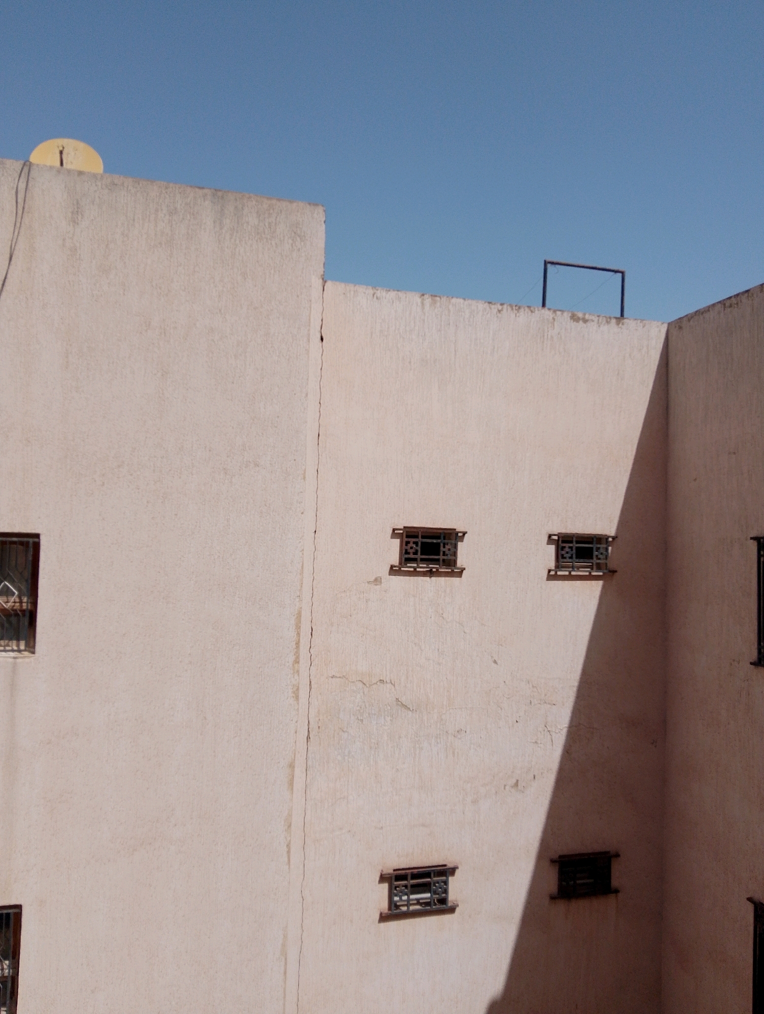 The image depicts a section of an exterior wall of a building in Fez, Morocco, under a clear blue sky. The wall is constructed from rough, textured material, painted in a light, faded pinkish-beige hue. A prominent vertical crack runs down the center-left portion of the wall.
Several small windows are visible, each fitted with decorative dark metal grilles. On the far left, a window is partially visible, with a hint of a curtain or blind. To the right of the crack, two similar windows are spaced horizontally. Further to the right, near the corner of the building, another window is present, and a portion of a fifth window is seen on the right edge of the frame. The light casts a distinct diagonal shadow across the wall, originating from an unseen architectural element.
Atop the wall, on the left side, a yellow satellite dish is mounted. In the background, above the right section of the wall, a simple, dark rectangular frame structure is visible against the sky. The overall impression is of an aged building facade in an urban setting, likely residential, with a functional yet somewhat weathered appearance.