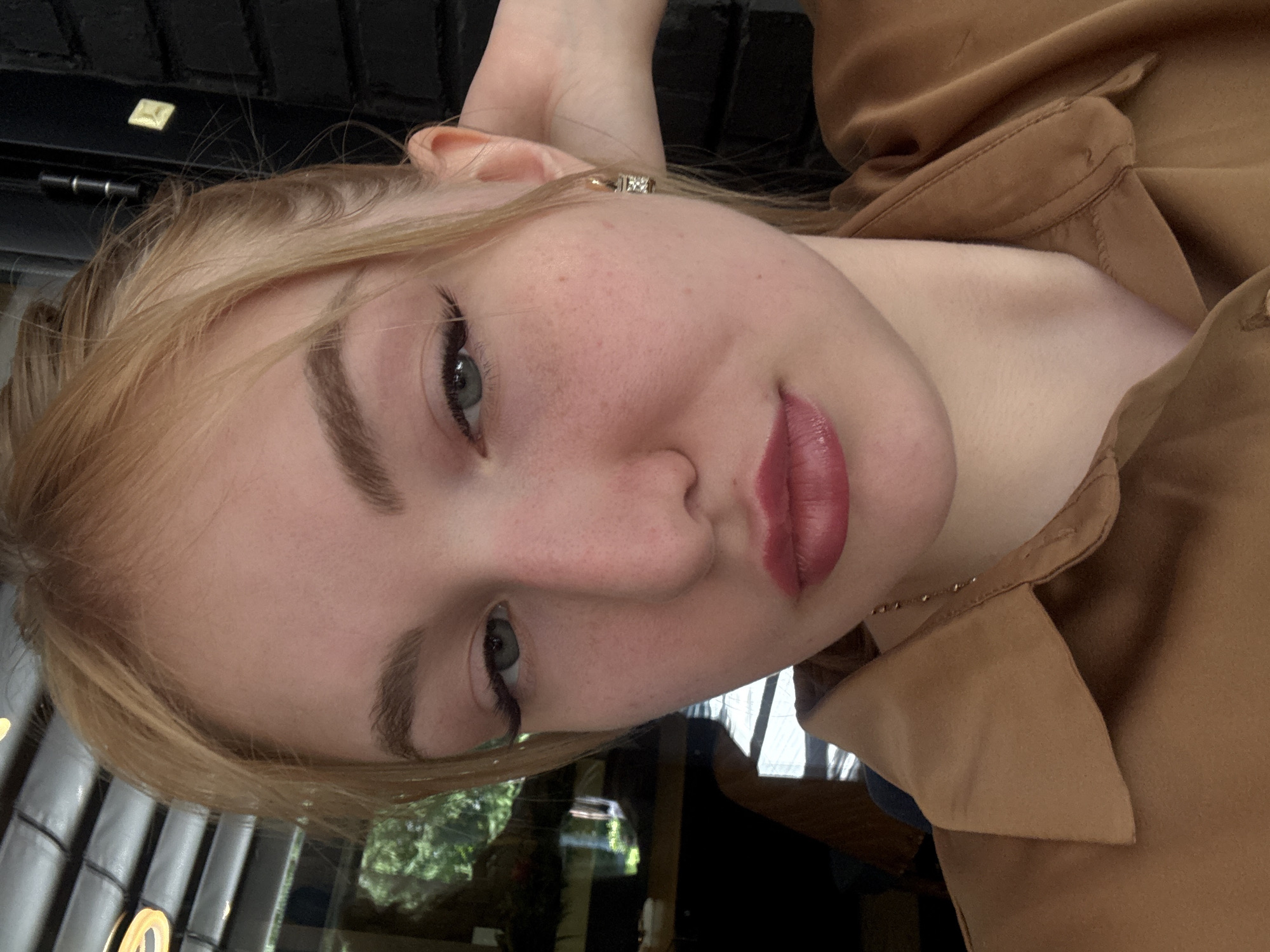 A young woman with blonde hair is taking a selfie. She is wearing a brown shirt with a collar and a necklace. She has on eyeliner and lipstick. Her right hand is behind her head. In the background, there is a window with trees outside and a brick wall. The image appears to be taken indoors, possibly in a cafe or restaurant.