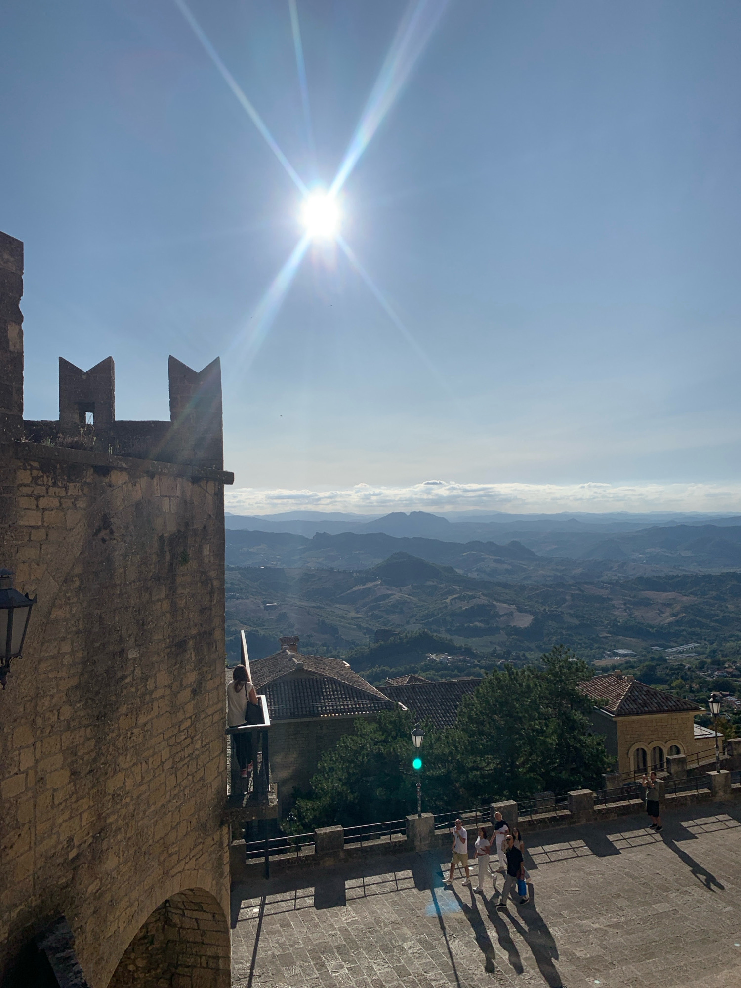 The image captures a sun-drenched vista from an elevated position in the City of San Marino, San Marino, likely from within the historic fortifications. In the foreground, a weathered stone battlement with crenellations runs along the left side, casting a sharp shadow. To the right, a paved courtyard area is visible, with a group of five individuals walking and observing the view. A street lamp with a green light is situated near the edge of the courtyard. Behind the courtyard, several terracotta-roofed buildings are clustered, suggesting a European old town. The background reveals a sprawling landscape of rolling hills and mountains fading into a hazy horizon, under a bright blue sky dominated by the sun, which creates a starburst effect.