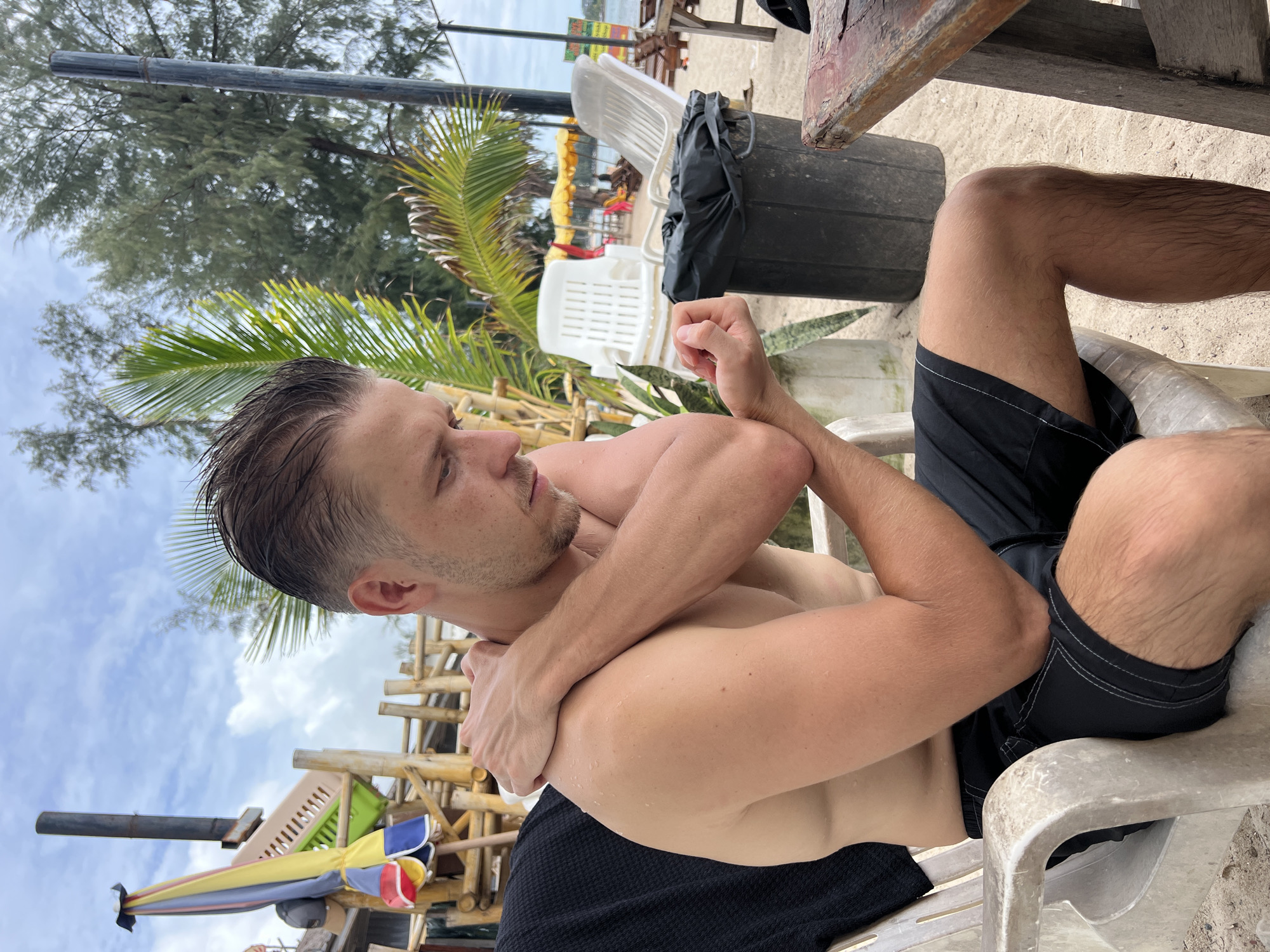A man with short brown hair, wearing black swim trunks, is seated on a white plastic chair on a sandy beach. He is holding his left arm across his chest with his right arm. The man appears to be looking down at his arm. In the background, there are palm trees, beach umbrellas, lounge chairs, and a trash can. The sky is overcast.